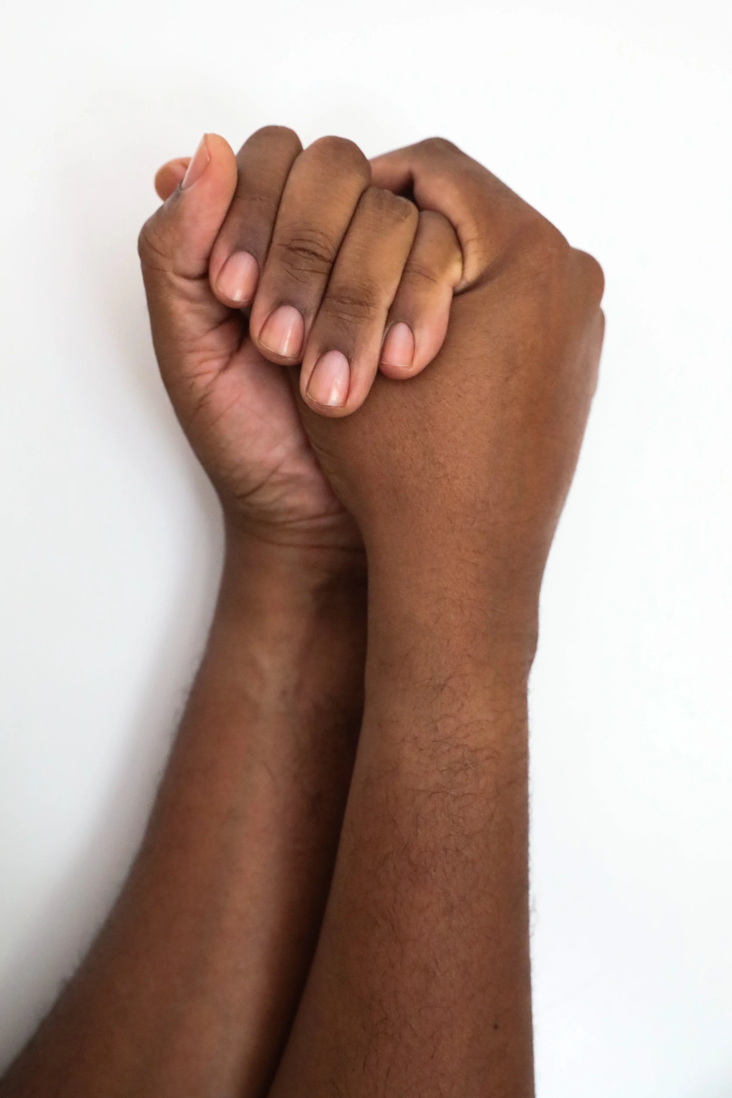 Two hands with richly hued,  medium dark skin tone and clean, clear nailbeds are clasped together against a plain white background.