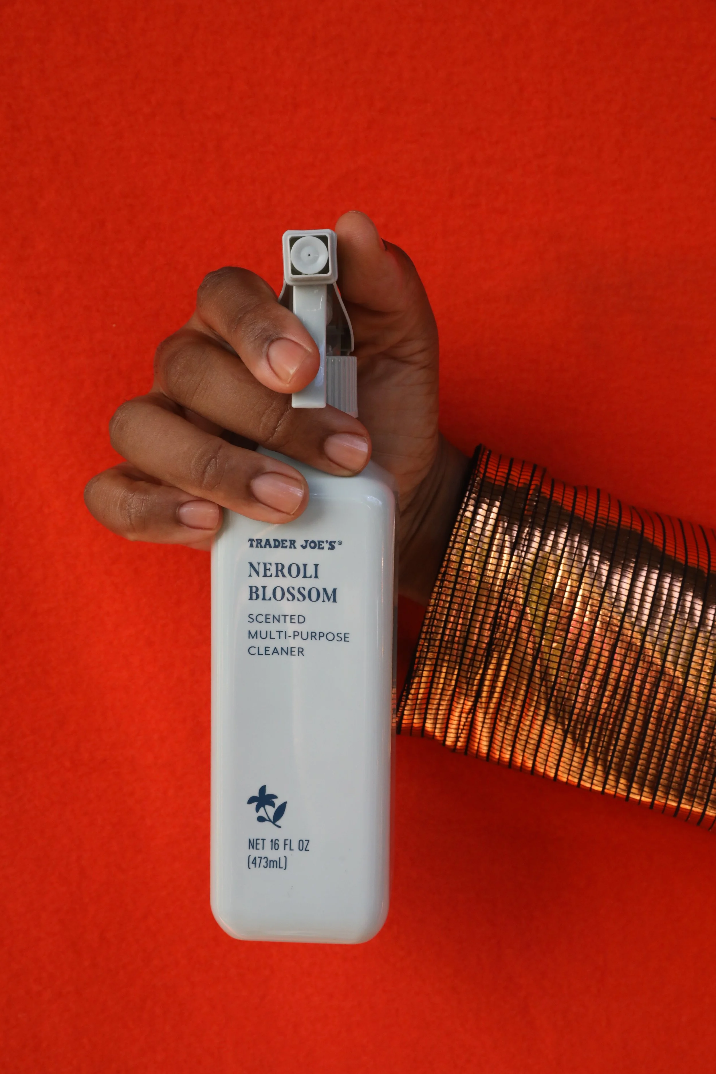 A richly hued, medium dark skin tone hand with clean, clear nailbeds holding a spray bottle of Trader Joe's  multi-purpose cleaner against a bright orange background  with part of a vibrantly metallic orange patterned sleeve visible.