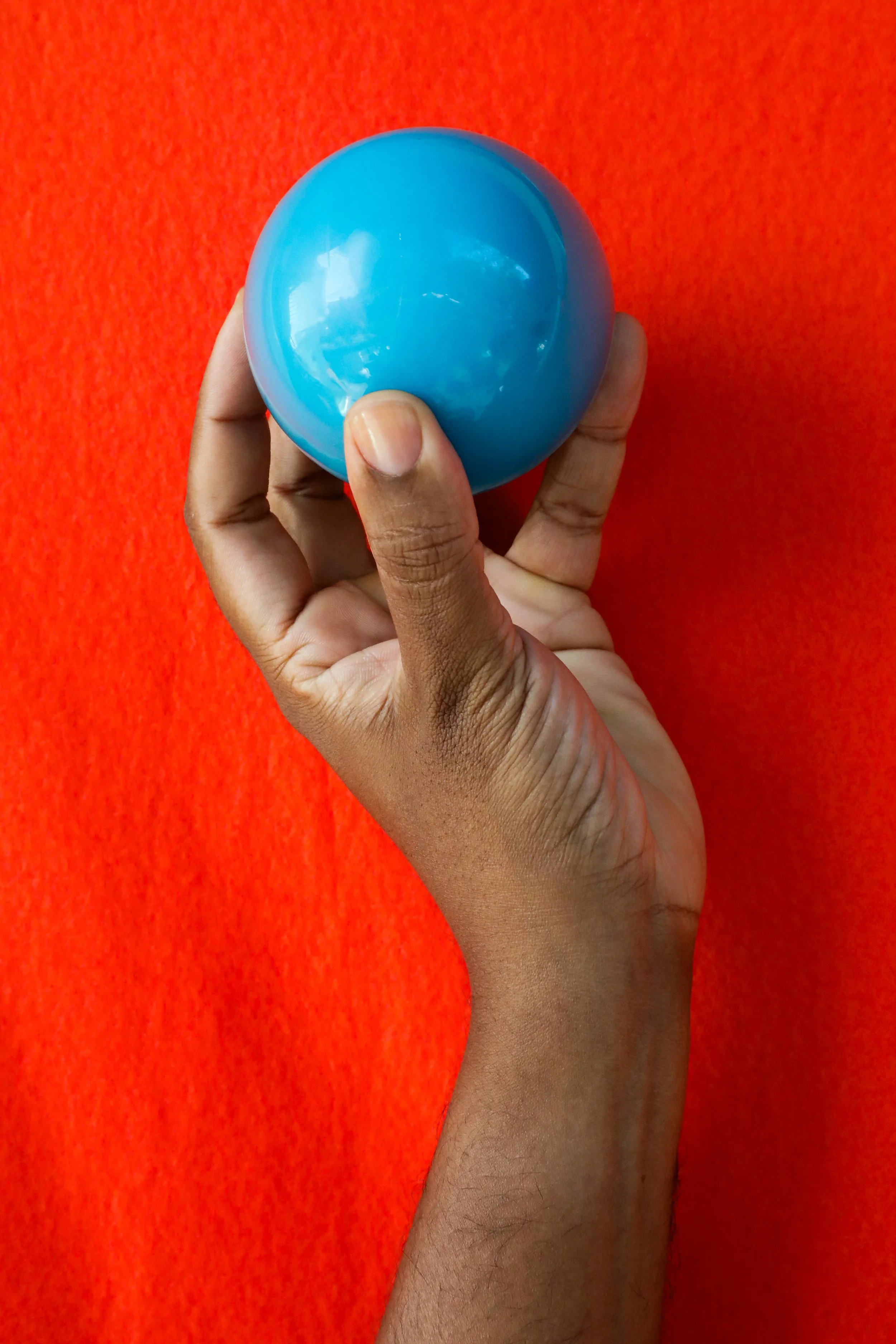 A richly hued, medium dark skin tone hand with clean, clear nailbeds holding a blue plastic ball against a bright orange background.