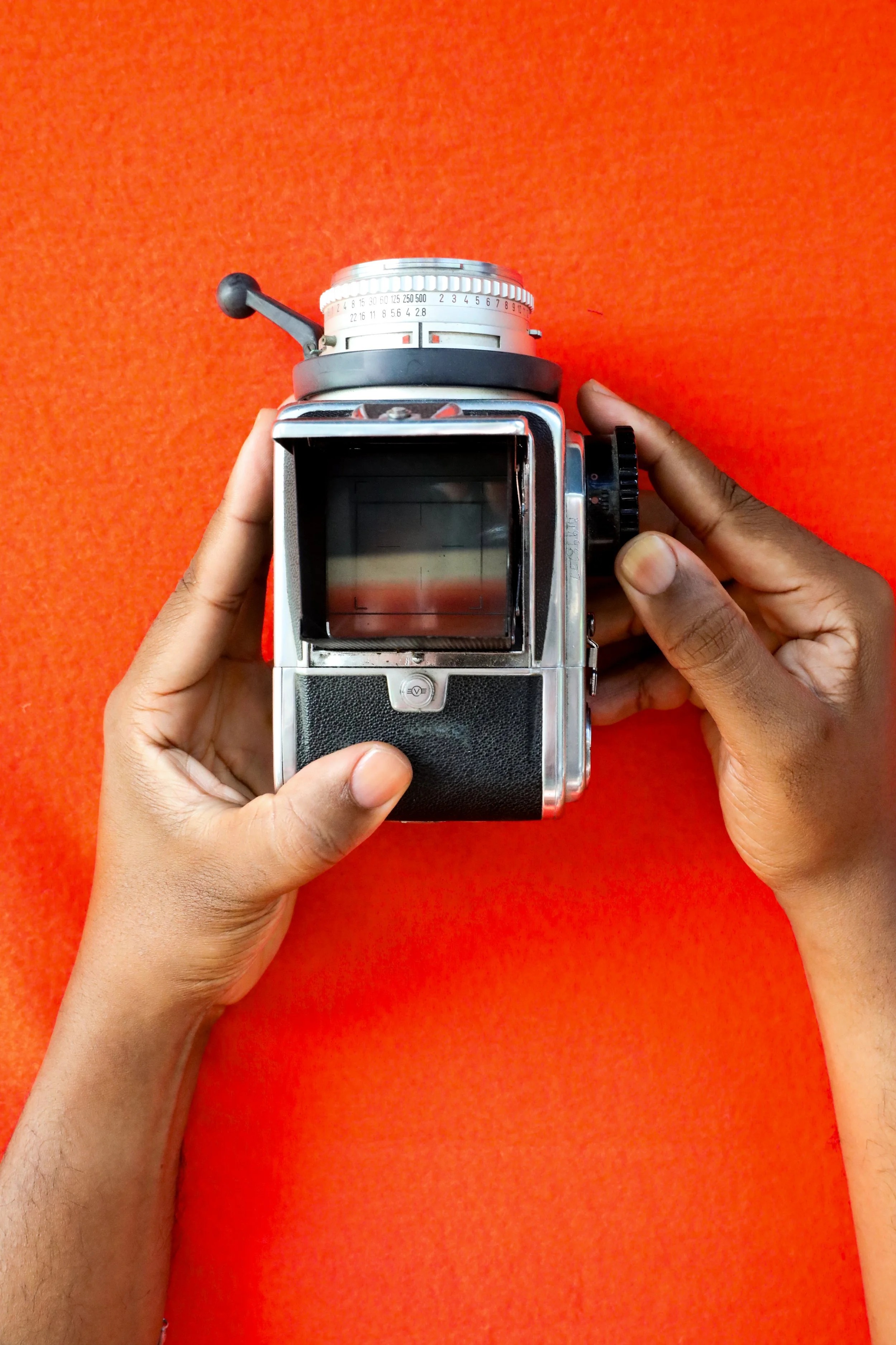 A person's richly hued, medium dark skin tone hands and clean, clear nailbeds holding an antique camera against a bright orange background.