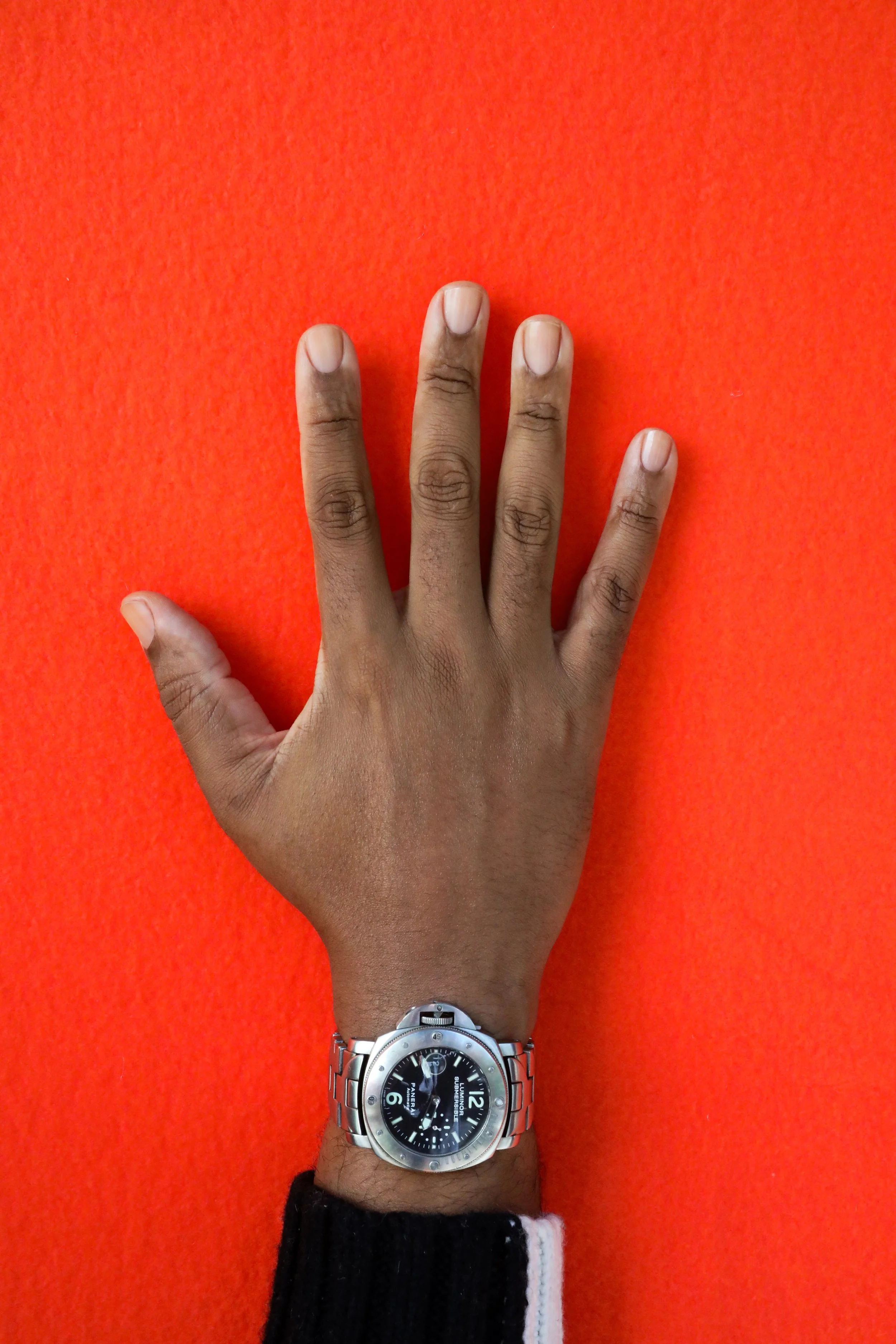 A person's richly hued, medium dark right hand with clean, clear nailbeds wearing a black, leather wristwatch with a silver frame and black and white wool sweater against an orange background.
