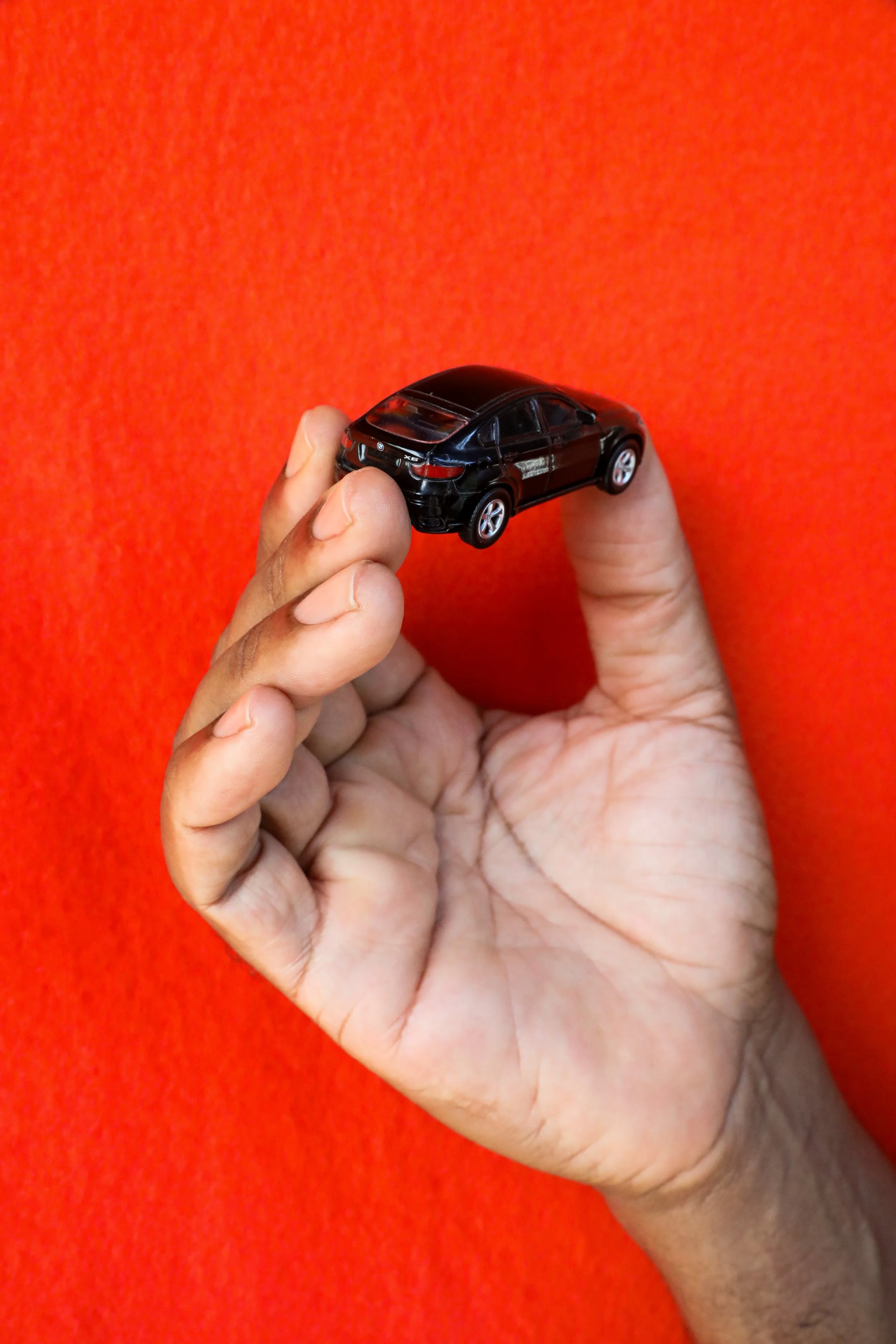 A richly hued, medium dark skin tone hand with clean, clear nailbeds holding a small toy BMW X6 SUV against a bright orange background.