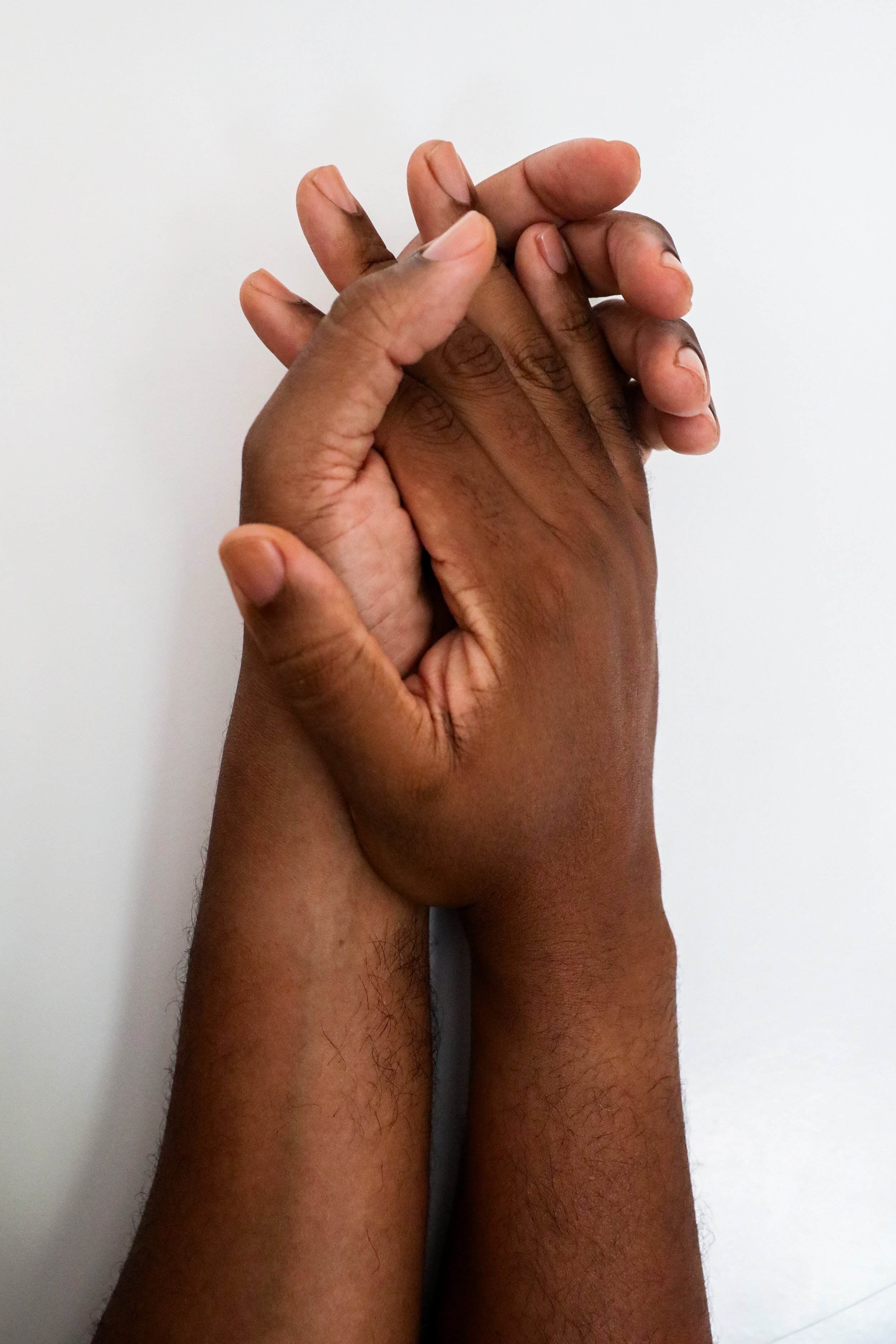 Two richly hued, medium dark hands with clean, clear nailbeds holding each other against a white background.