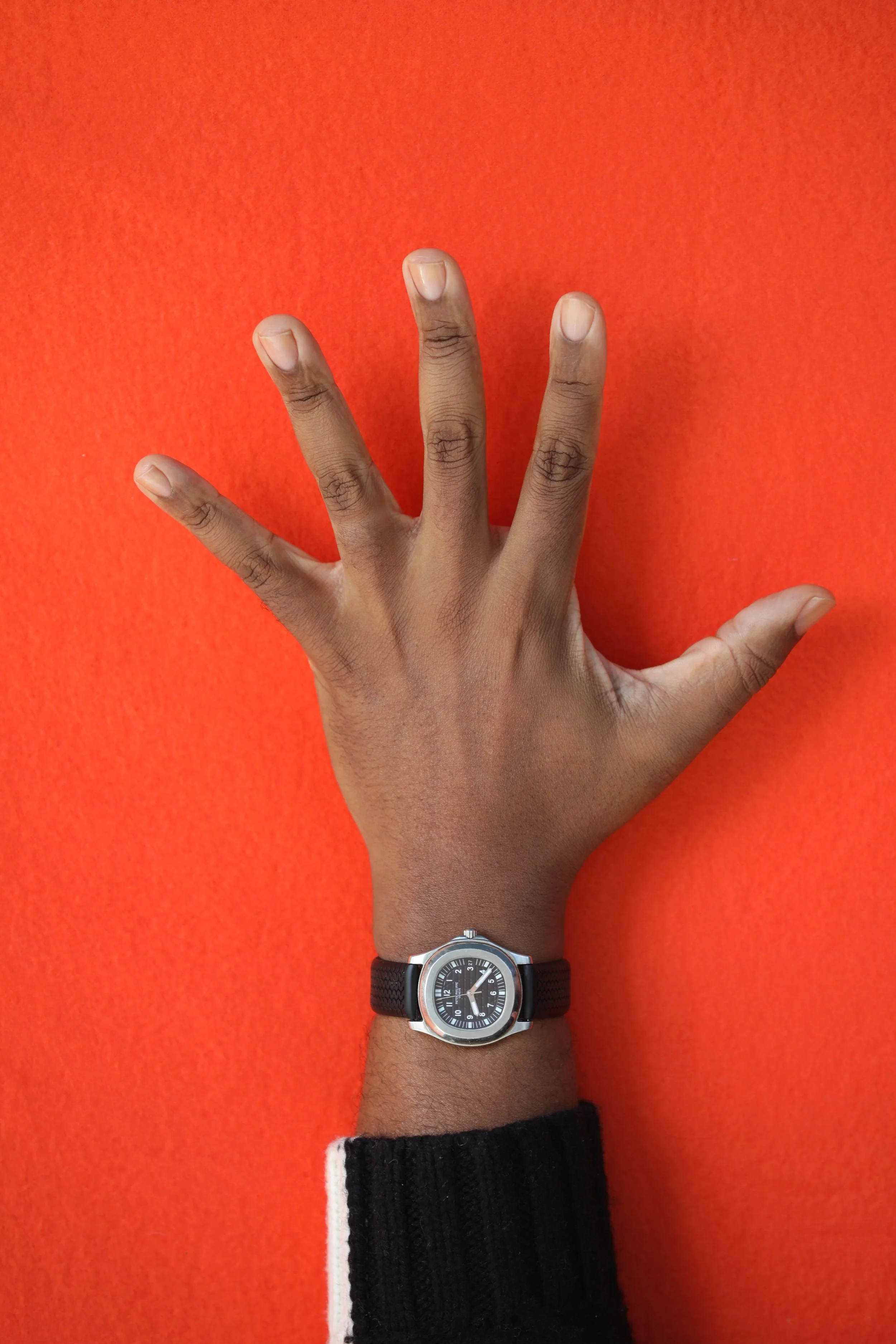 A person's richly hued, medium dark left hand with clean, clear nailbeds wearing a black, leather wristwatch with a silver frame and black and white wool sweater against an orange background.