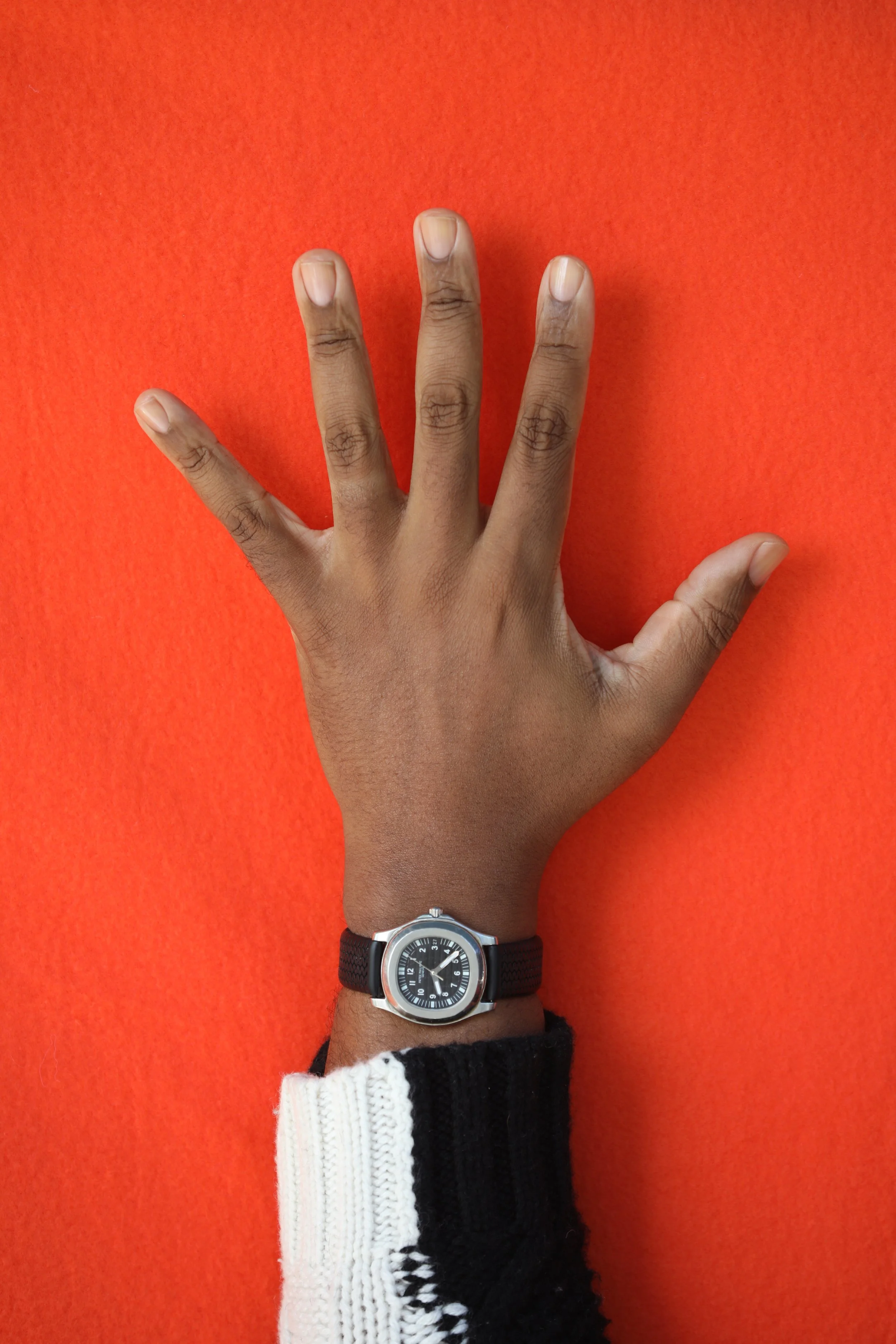 A person's richly hued, medium dark left hand with clean, clear nailbeds wearing a black, leather wristwatch with a silver frame and black and white wool sweater against an orange background.