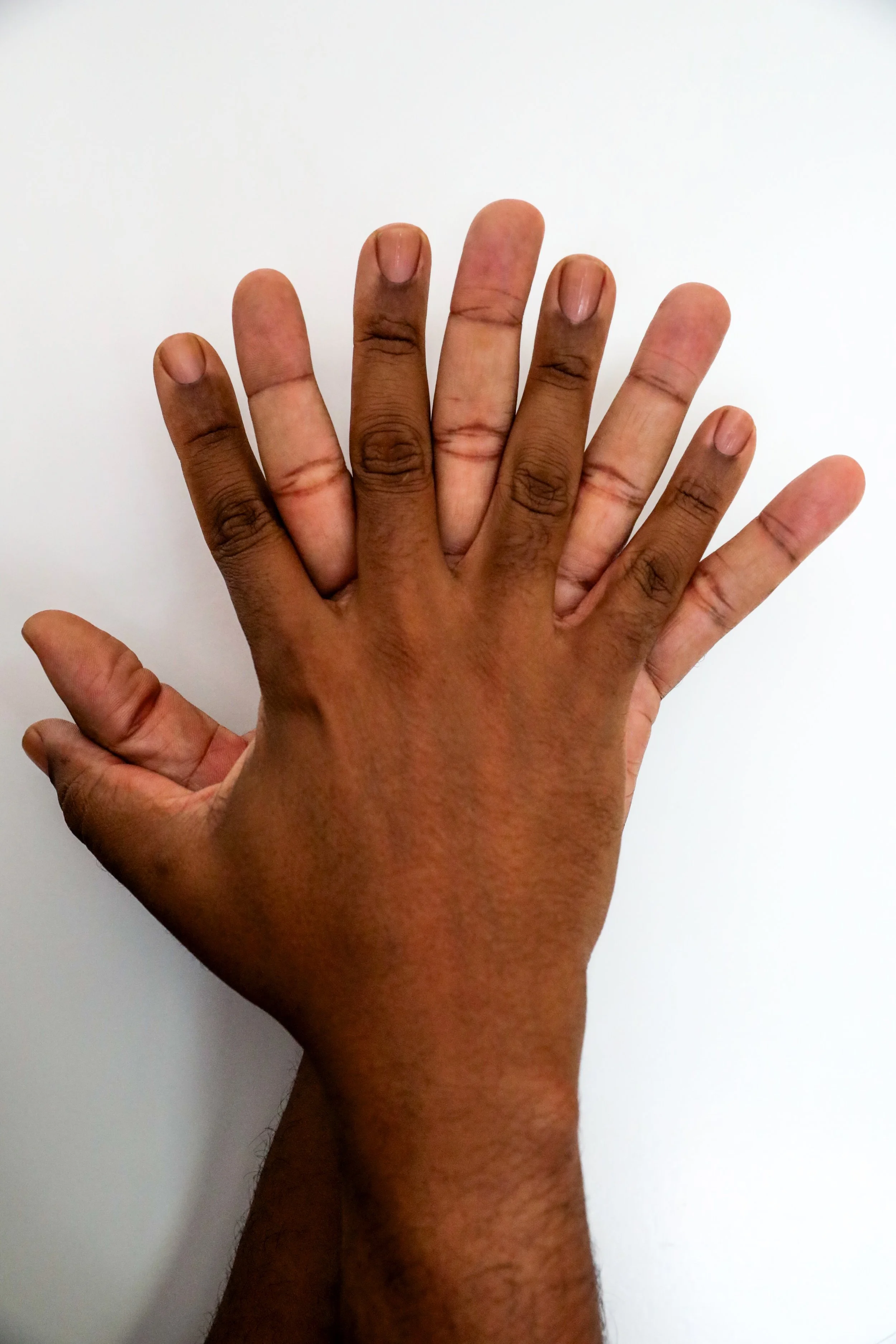 Two richly hued, medium dark hands with clean, clear nailbeds with fingers intertwined against a plain white background.