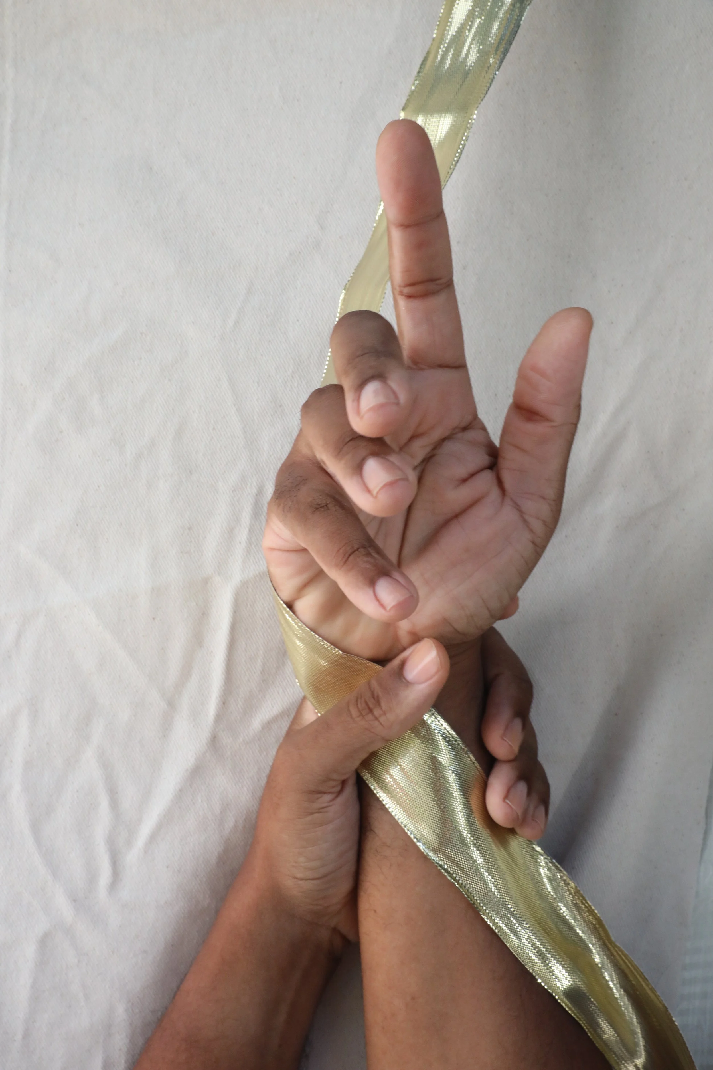Two hands of richly hued, medium dark skin tone with clean, clear nailbeds holding a piece of gold ribbon against a plain white background.