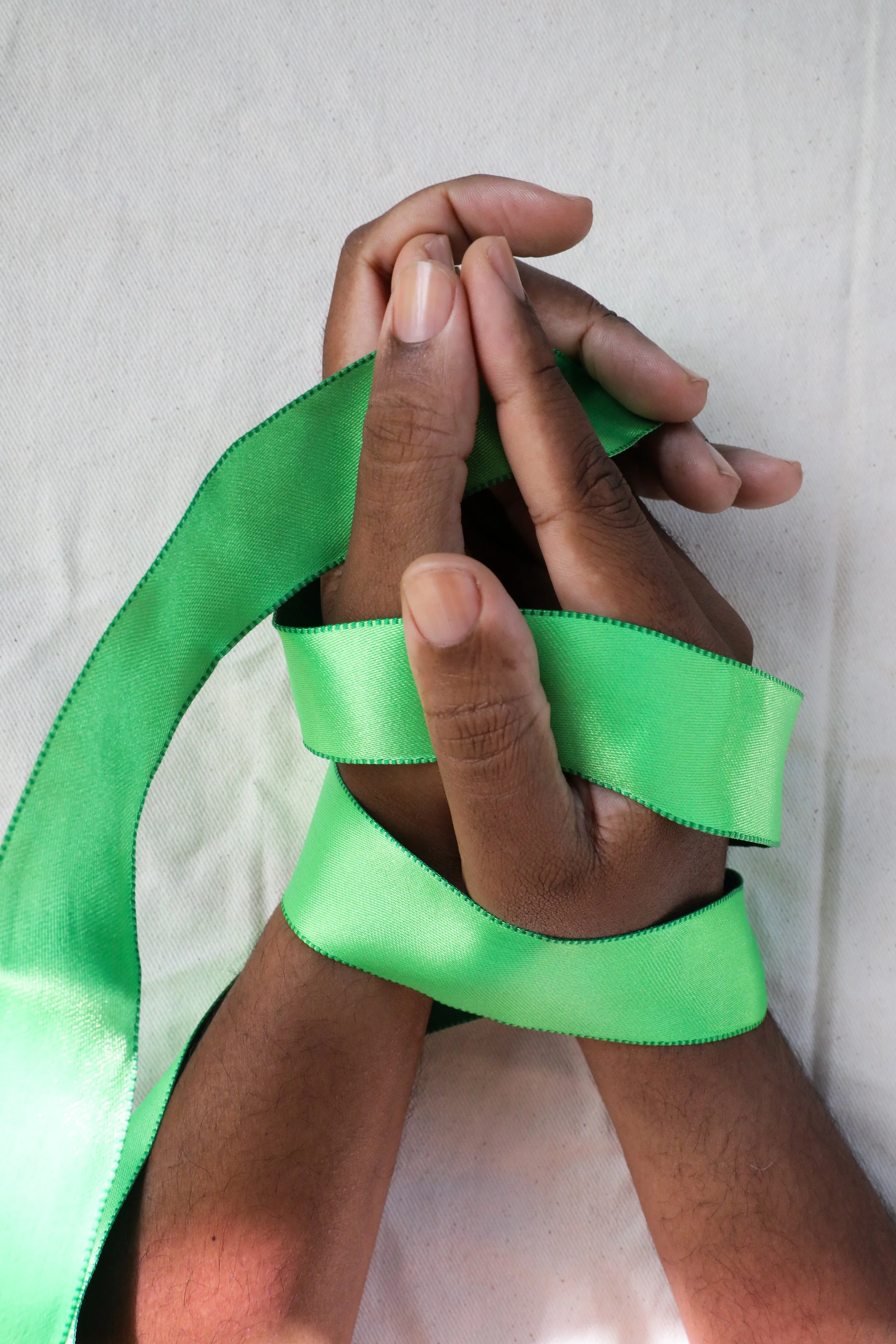 Two hands of richly hued, medium dark skin tone with clean, clear nailbeds holding a piece of green ribbon against a plain white background.