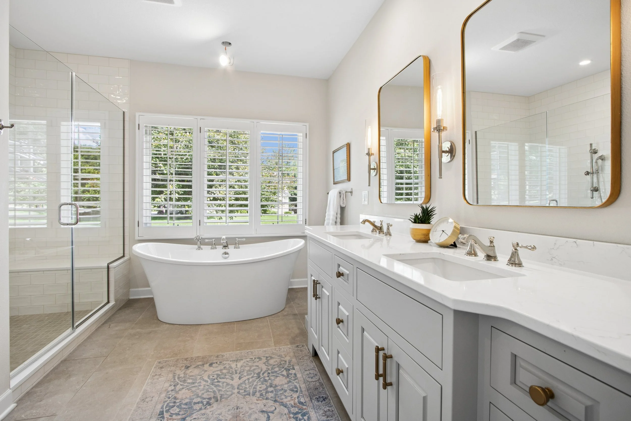 Bright bathroom with a large window, white vanity with dual sinks, mirrors, a freestanding bathtub, and a walk-in shower with glass door.
