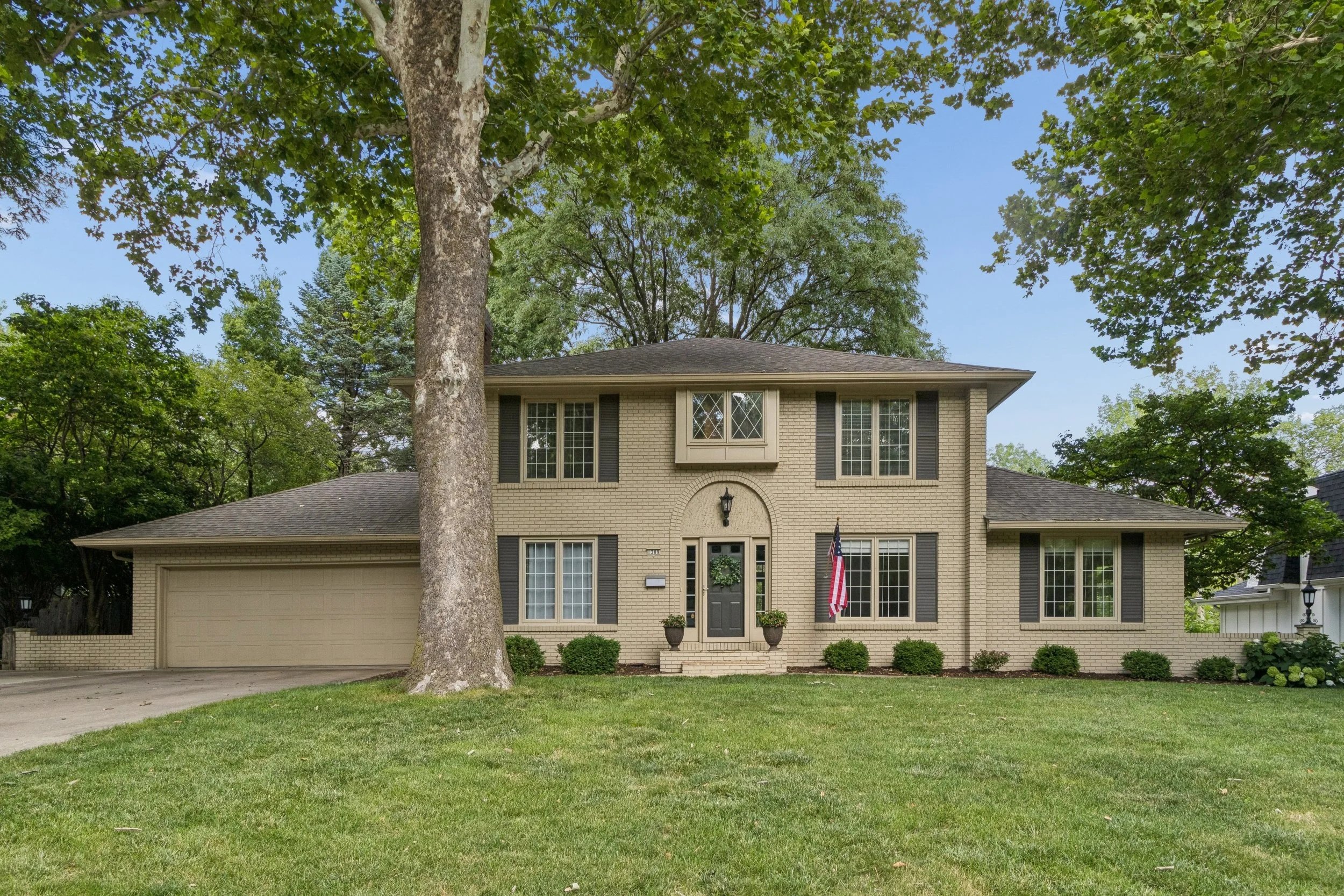 A two-story house with a brick facade, a dark gray front door with a wreath, an American flag hanging outside, and white-framed windows, surrounded by trees and a well-manicured lawn.