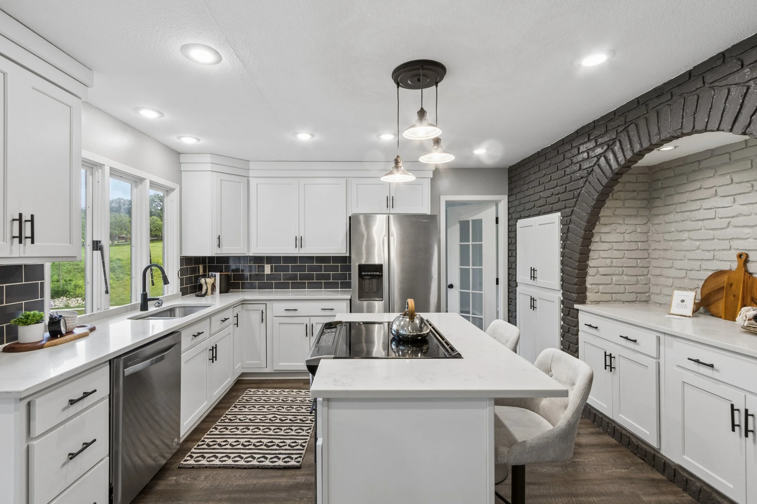 Modern kitchen with white cabinets, black subway tile backsplash, stainless steel appliances, and a white island with chairs. There is a window with a view of greenery outside and a black brick accent wall with an arch.