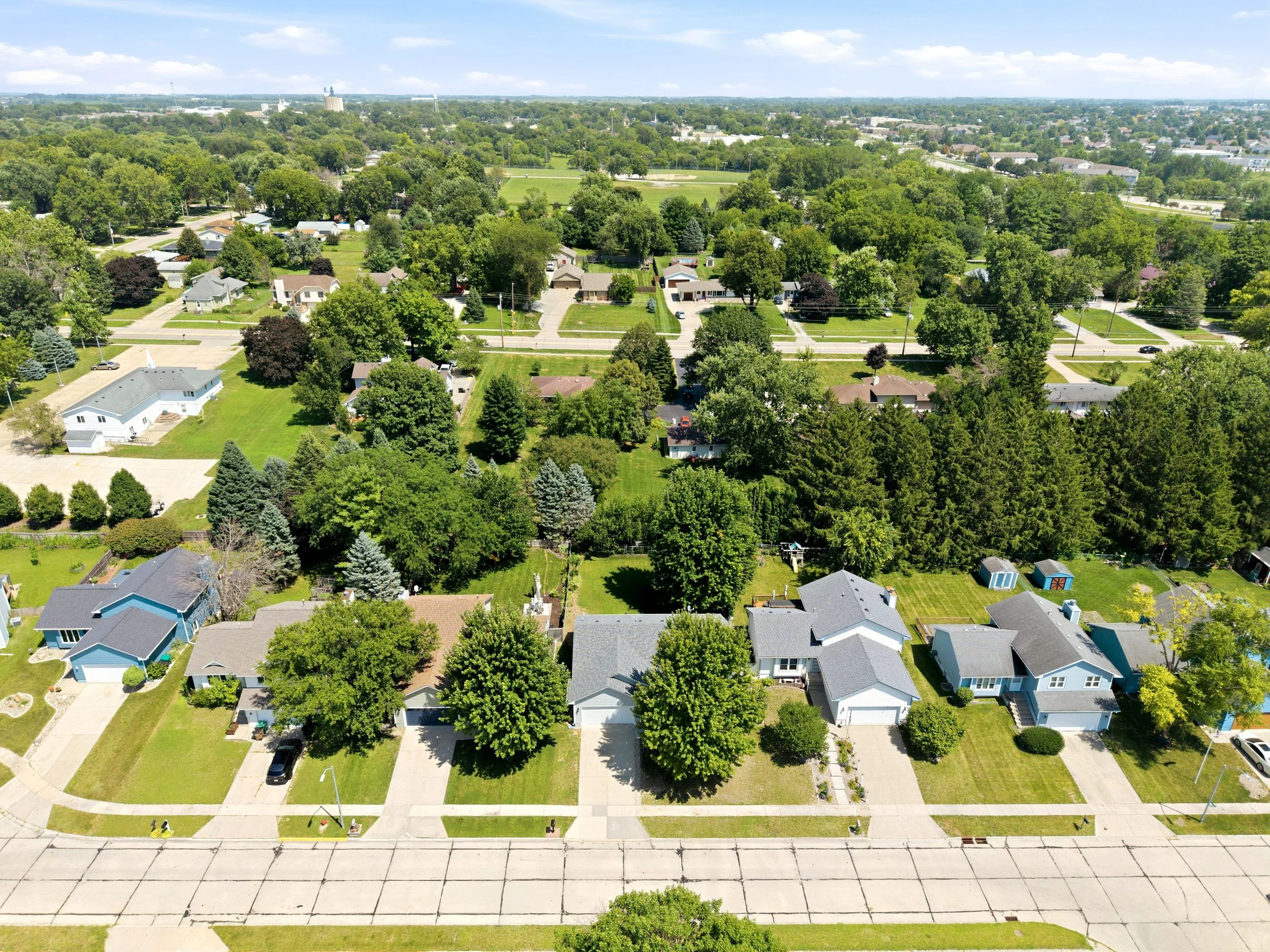 Aerial view of a suburban neighborhood with houses, lush green trees, and a park in the background, under a partly cloudy sky.