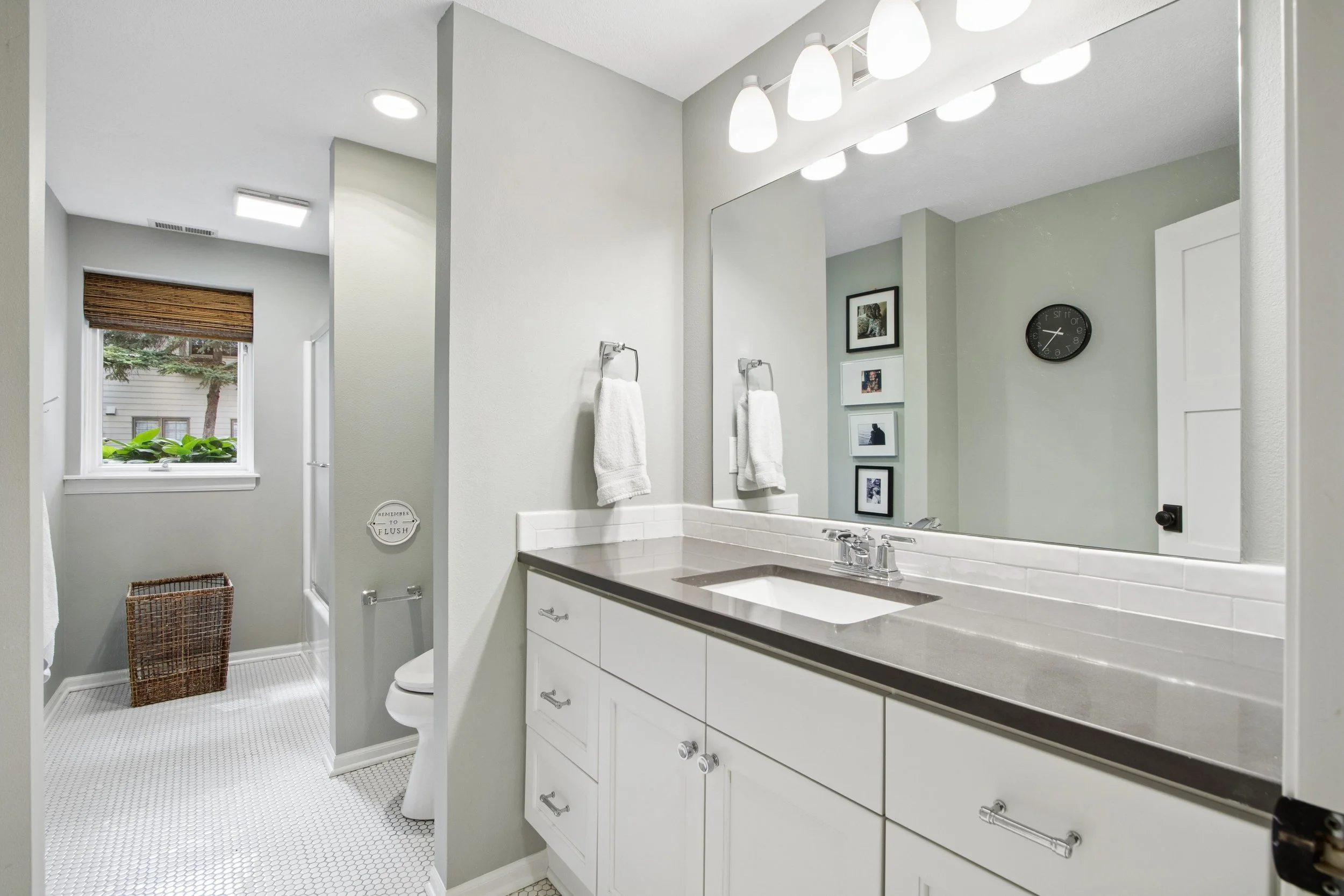Bathroom with a large mirror, white vanity with a gray countertop, framed pictures on the wall, window with brown blinds, and a tiled floor.