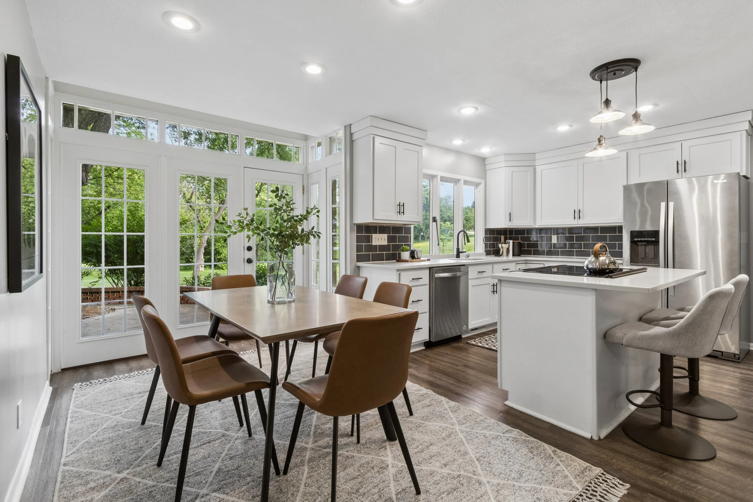 Bright kitchen with white cabinets, black backsplash, stainless steel appliances, wooden dining table with brown chairs, large windows and glass door showing green trees outside, ceiling lights, and a plant on the table.