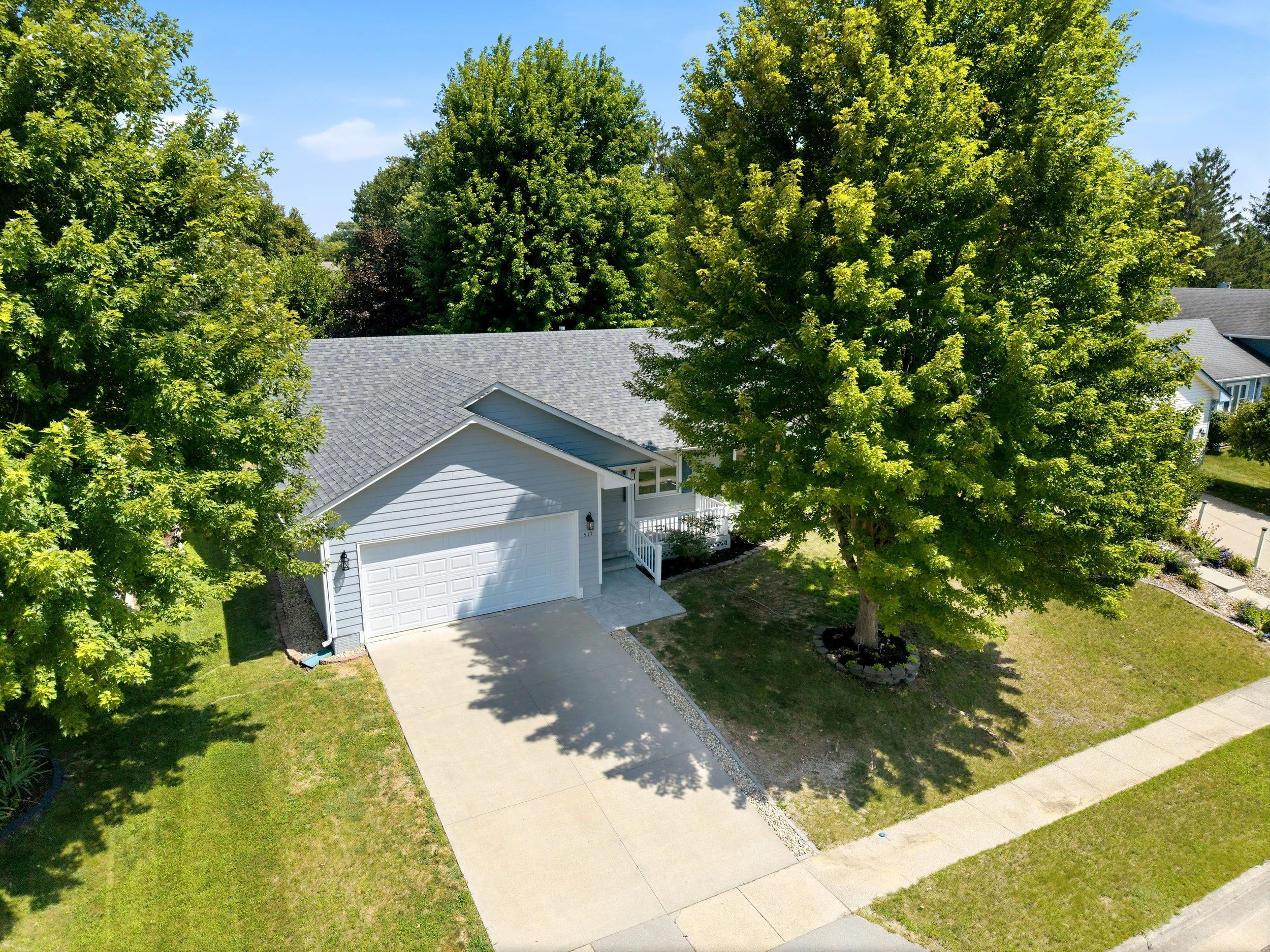 Aerial view of a suburban house with a gray roof, white siding, a driveway, and a front yard lush with green trees and grass, under a bright blue sky.