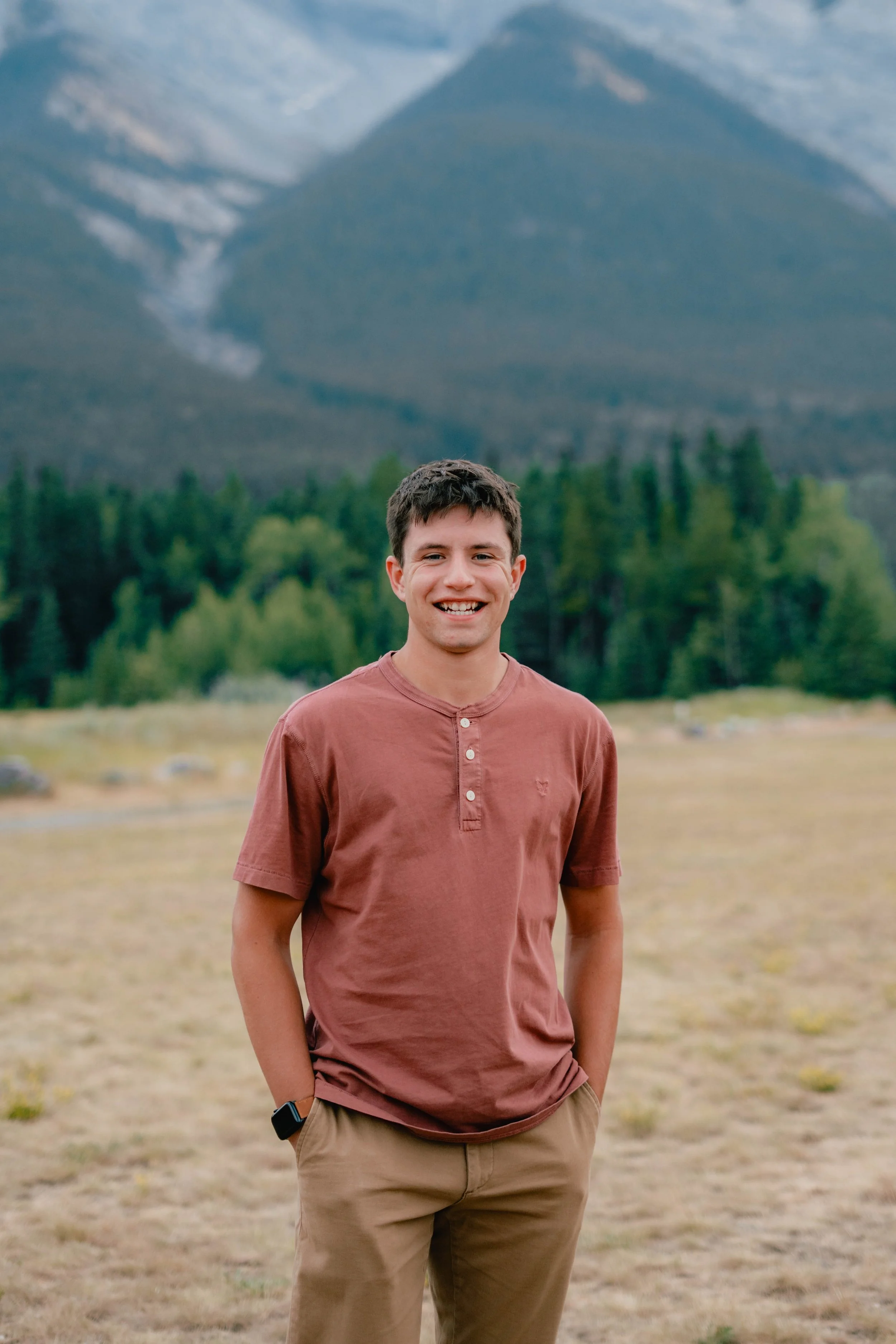 A young man standing outdoors in front of a mountain with snow-capped peaks and a forested area, smiling at the camera.