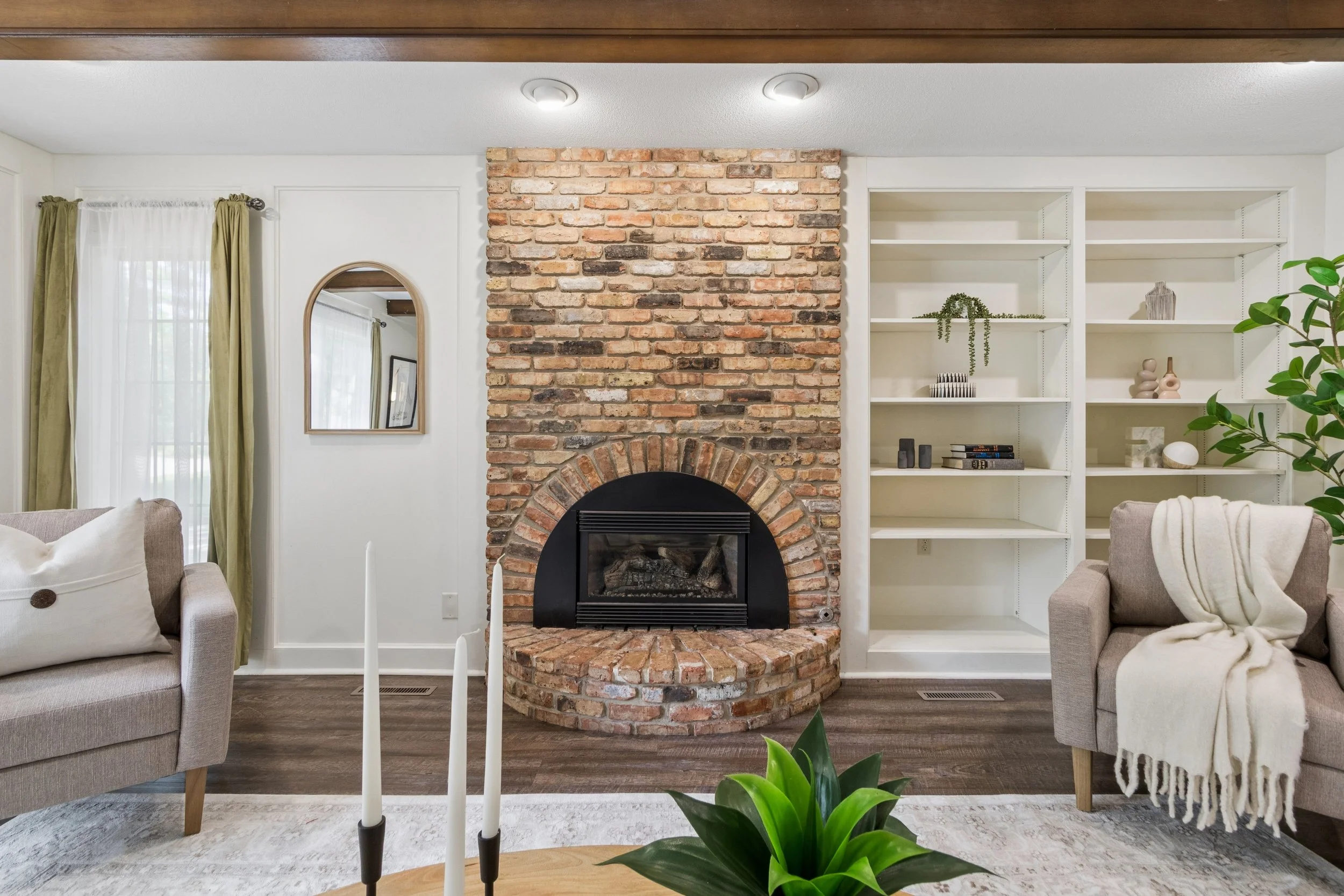 Living room with brick fireplace, two beige sofas, white bookshelf with decorative items, green plants, and a window with green curtains.