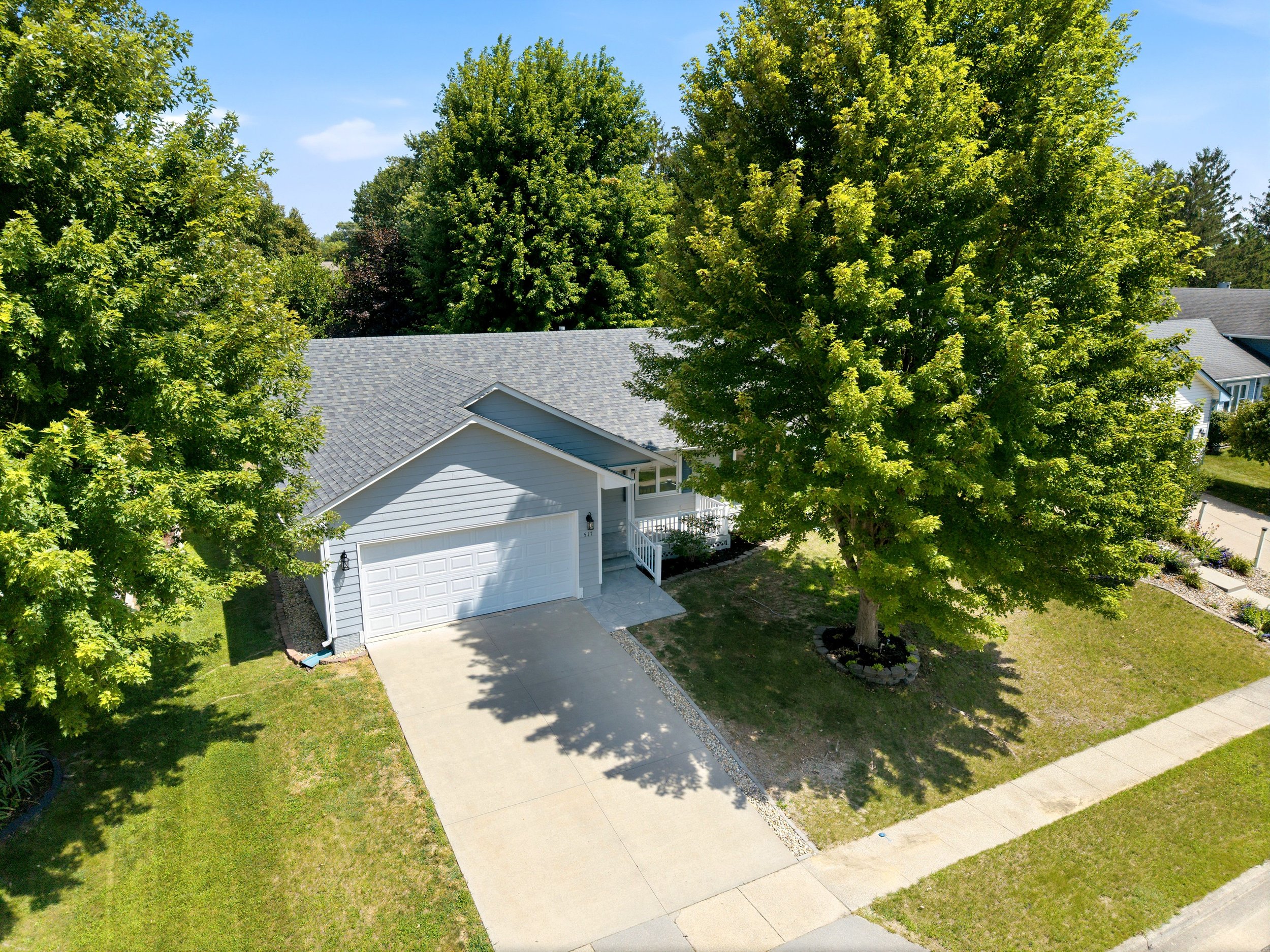 Aerial view of a residential house surrounded by greenery, including large trees and a grassy lawn, with a concrete driveway and a white garage door.