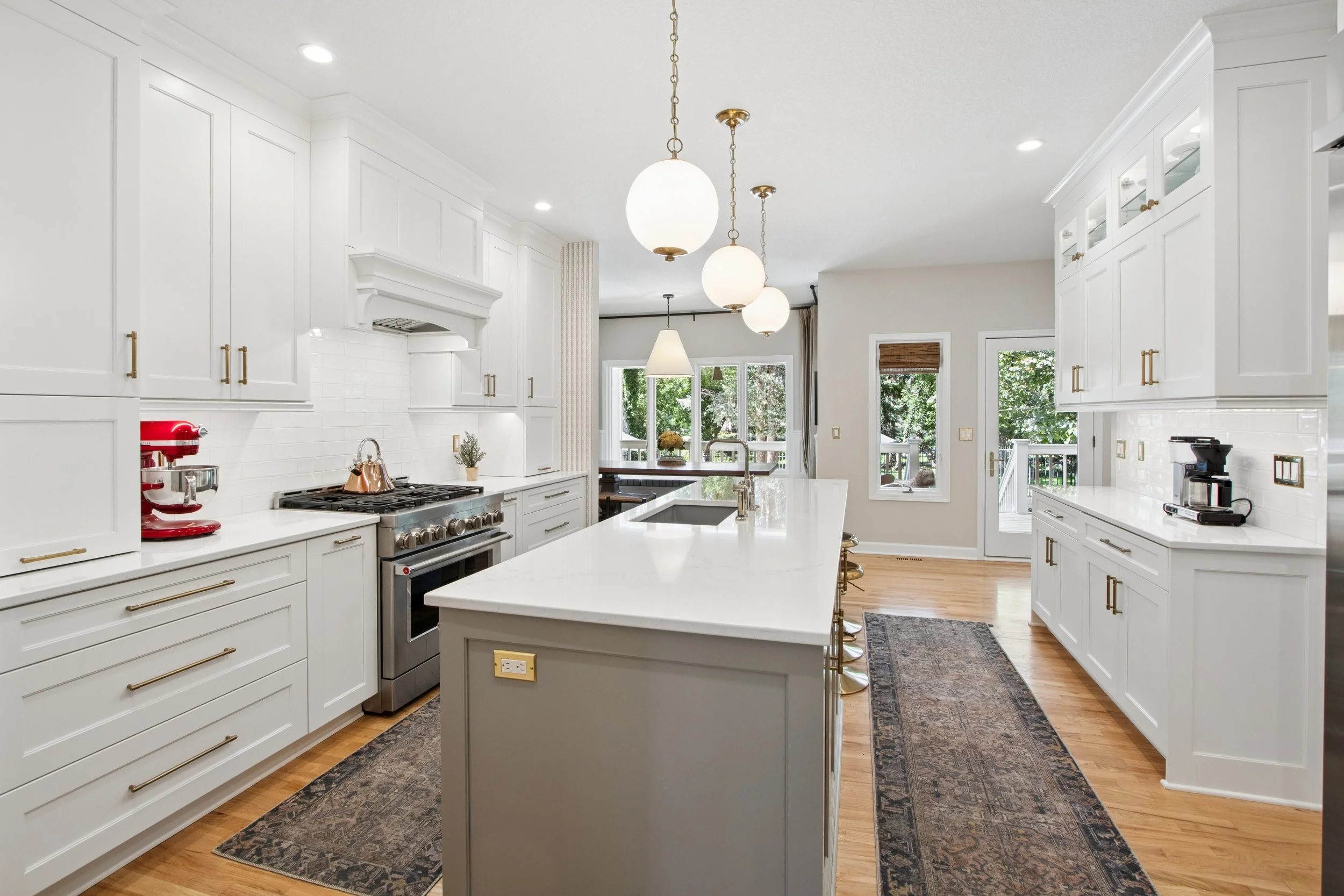 Bright, modern white kitchen with pendant lights, a large island, stainless steel appliances, and natural light from windows and a door leading outside.