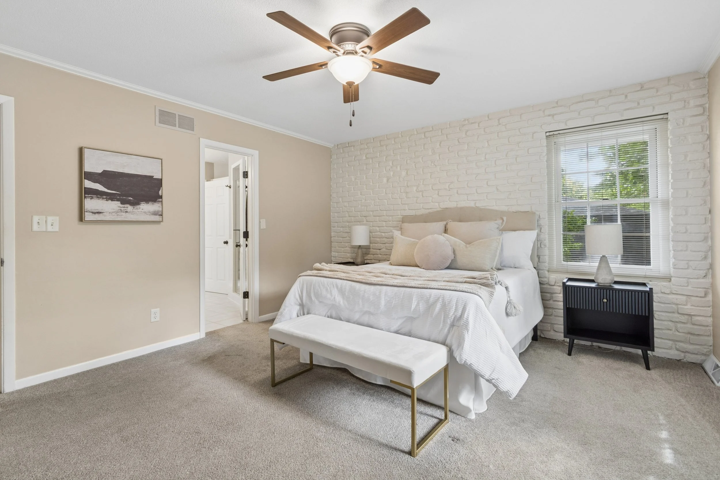 Bedroom with white brick accent wall, beige walls, beige upholstered bed with cream bedding and neutral pillows, black nightstand with lamp, window with blinds, and ceiling fan with wooden blades.