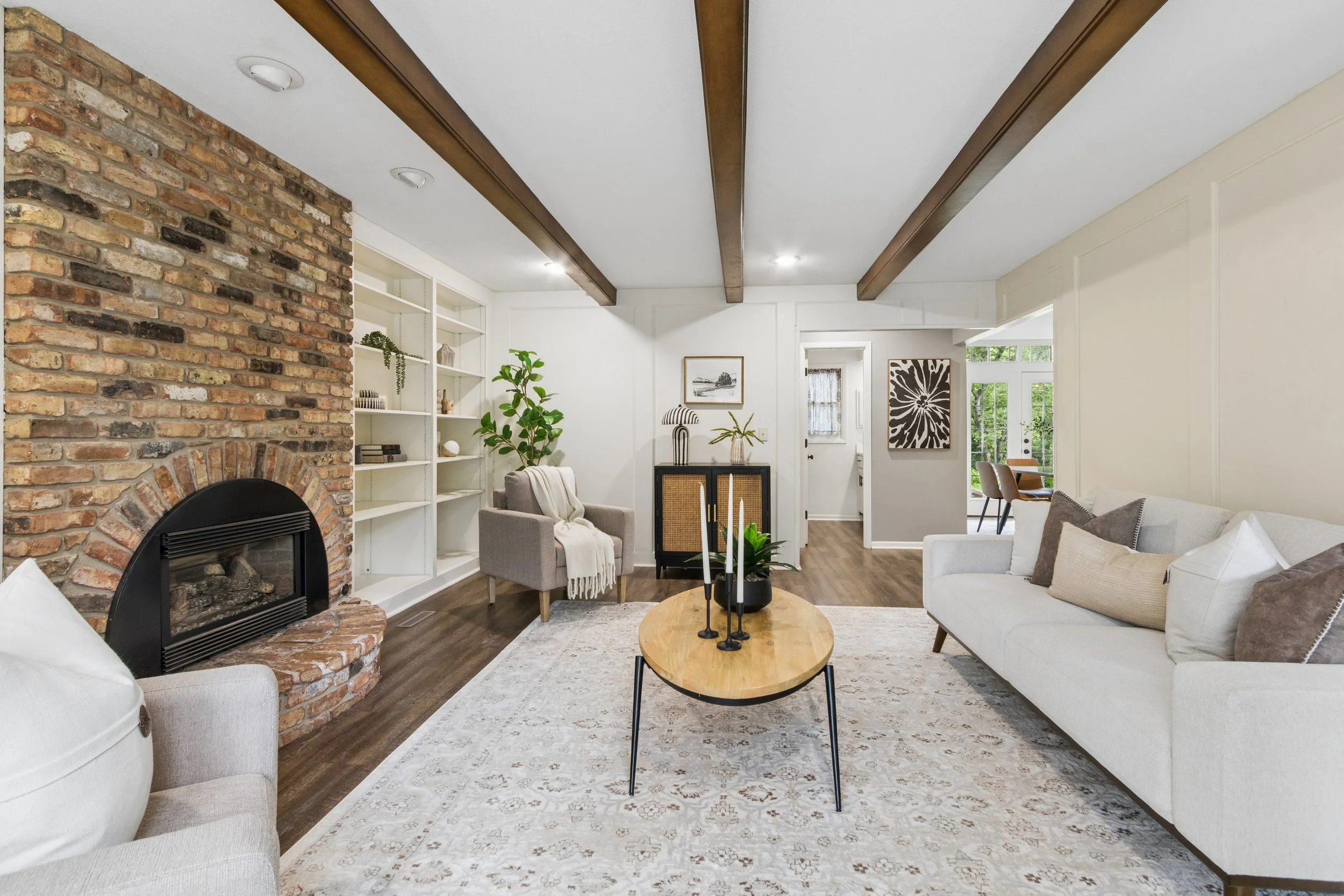 A cozy living room with a brick fireplace, white bookshelf, gray armchair, white sofa, and wooden coffee table. Decor includes a potted plant, lamps, and framed artwork, with a natural light coming from windows.