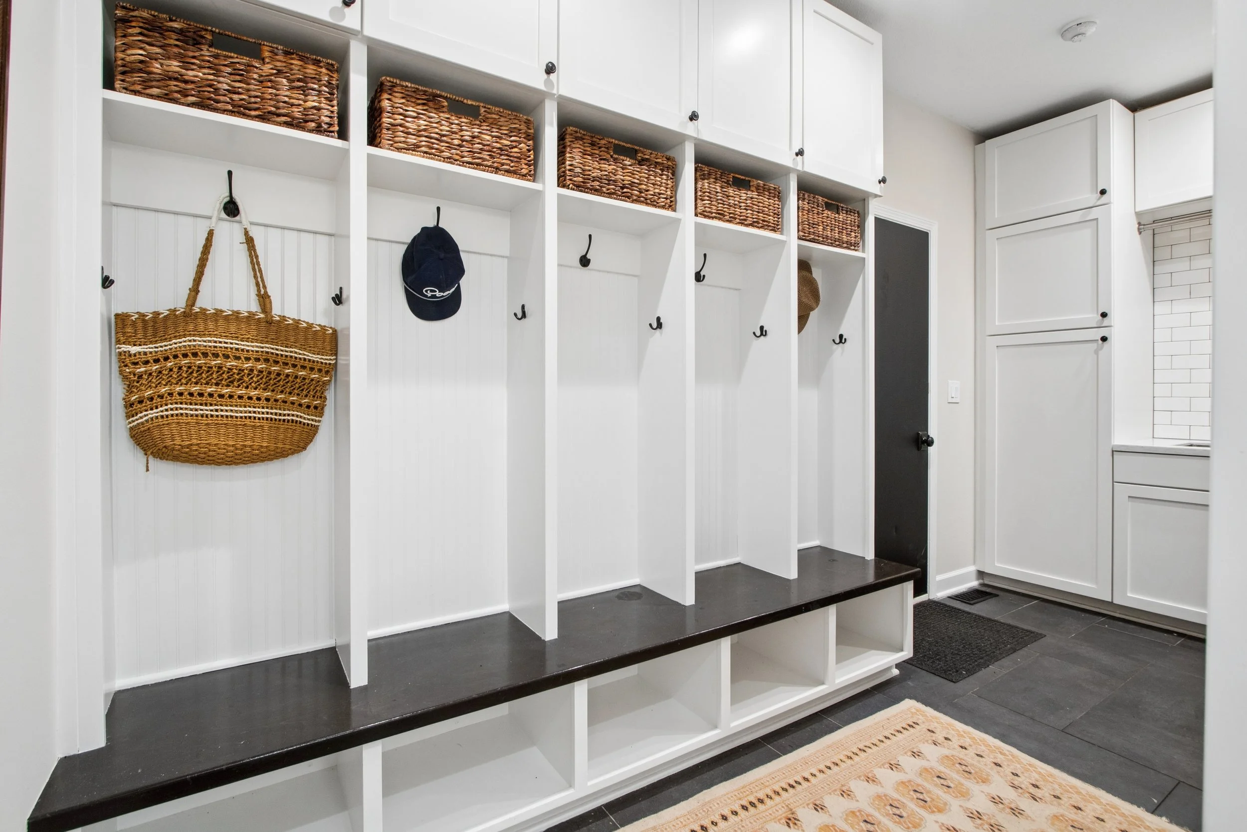 Entryway with white built-in storage and hooks, baskets on top, a woven bag hanging, a dark door, and a small dining or living area rug.