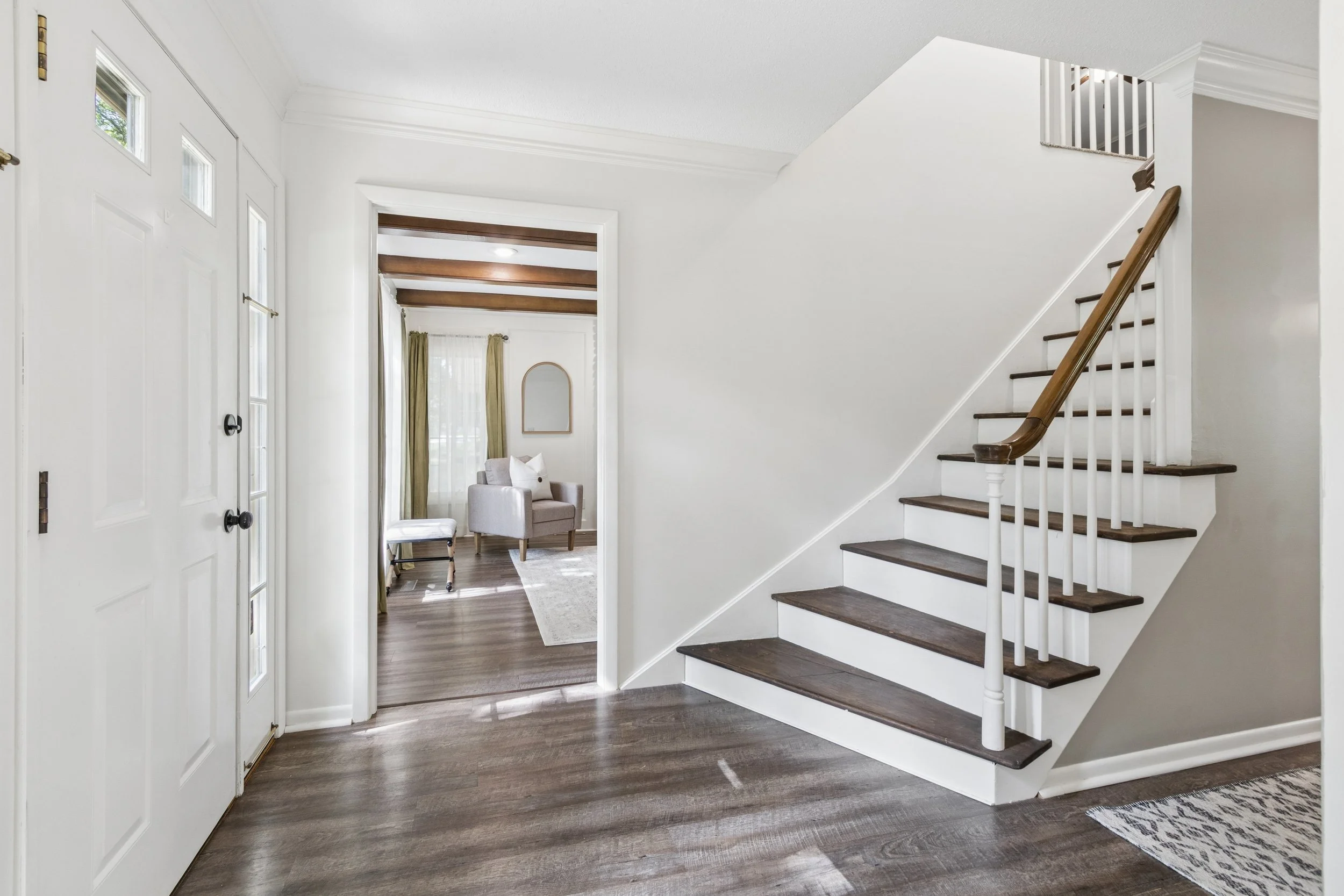 Foyer with dark wood flooring, white walls, staircase with dark wood steps and white banister, front door with glass panels, living room with beige armchairs and window with curtains visible in background.
