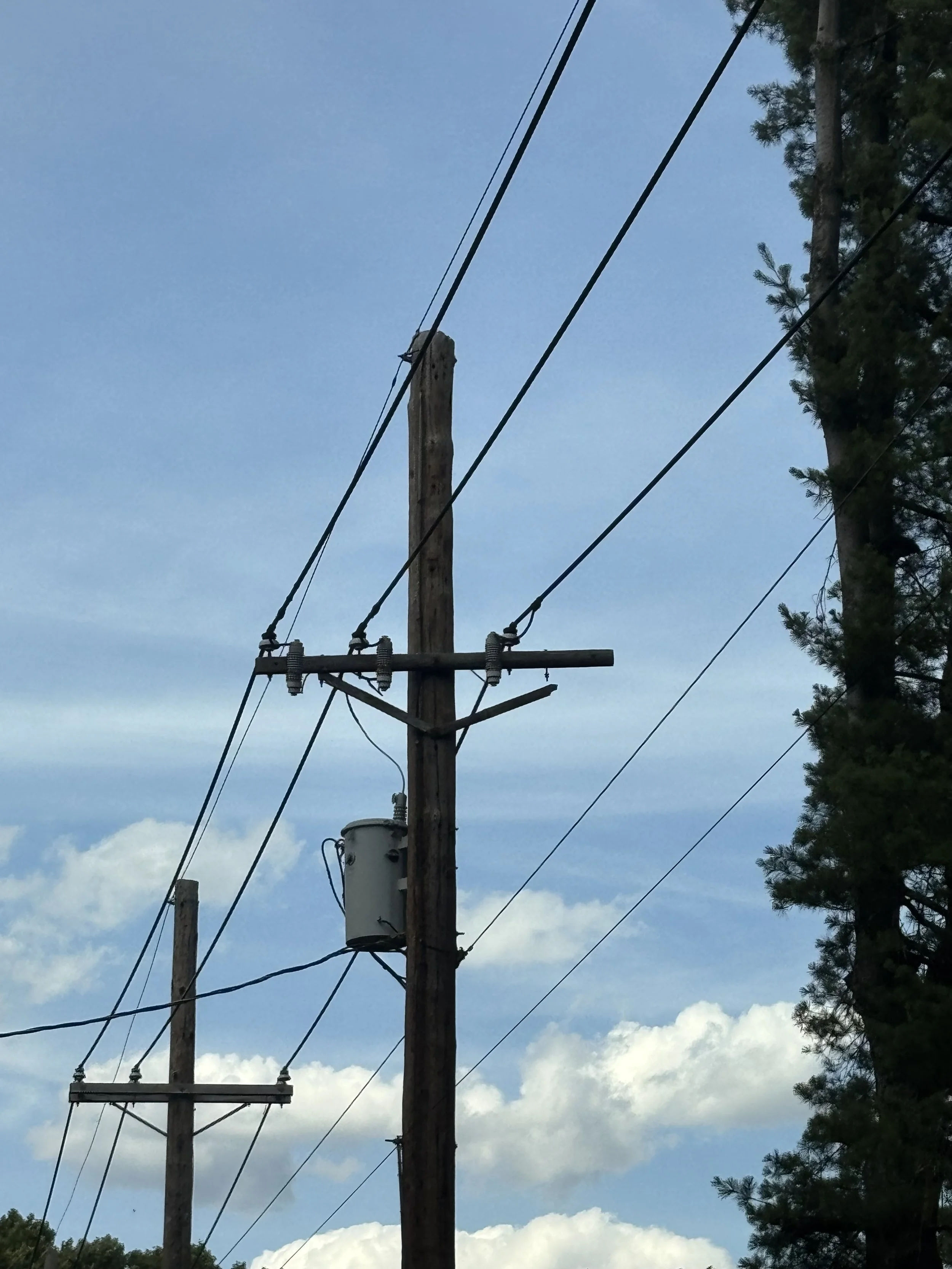 A wooden utility pole with power lines and a transformer, surrounded by trees and clouds in a blue sky.
