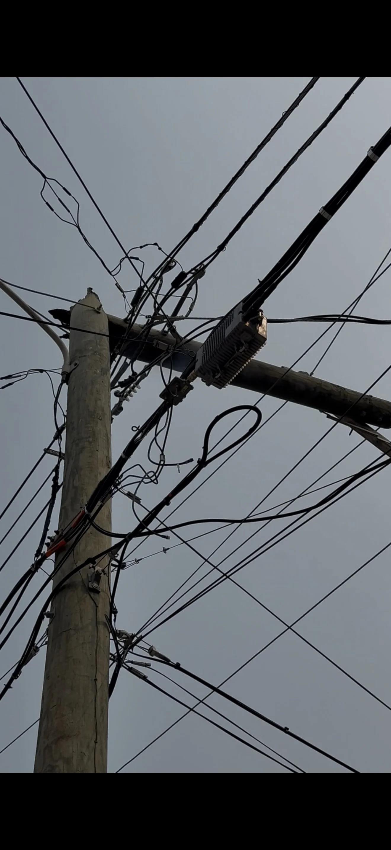 A close-up view of a wooden utility pole with multiple black power lines and wires attached, set against a cloudy sky.
