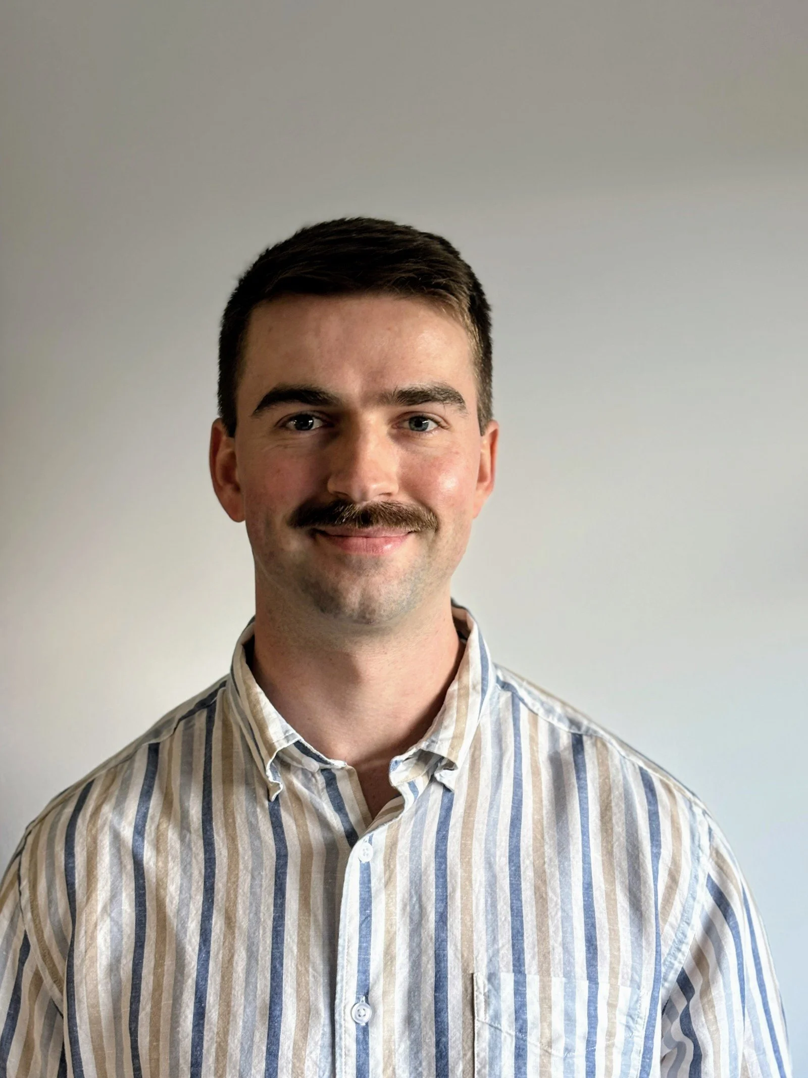 Portrait of a smiling young man with short, dark hair and a mustache, wearing a beige, white, and blue striped button-up shirt, standing against a plain, light-colored background.