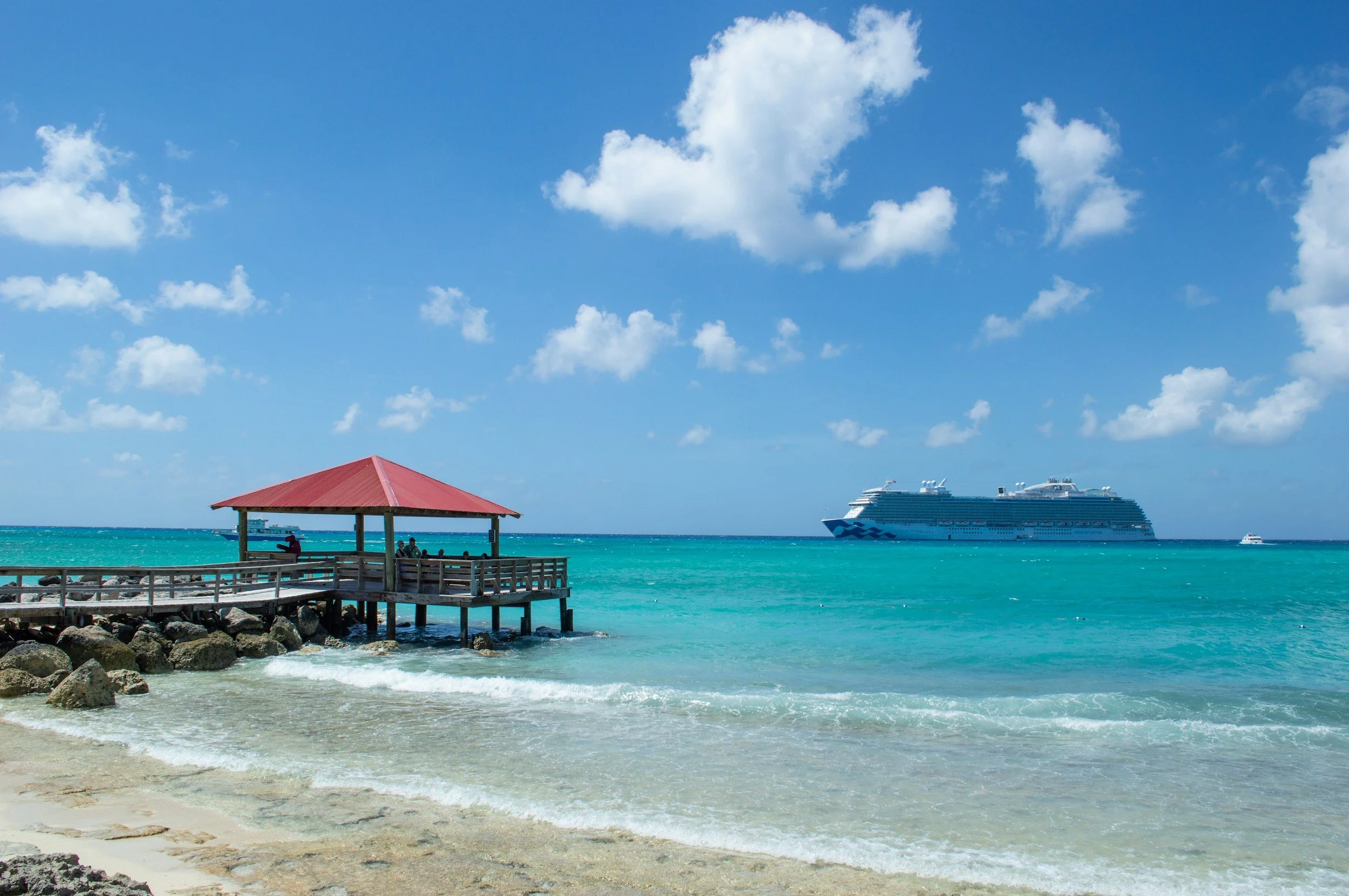 A tropical beach with turquoise water, a wooden pier with a red roof, a cruise ship in the distance, and a partly cloudy blue sky.