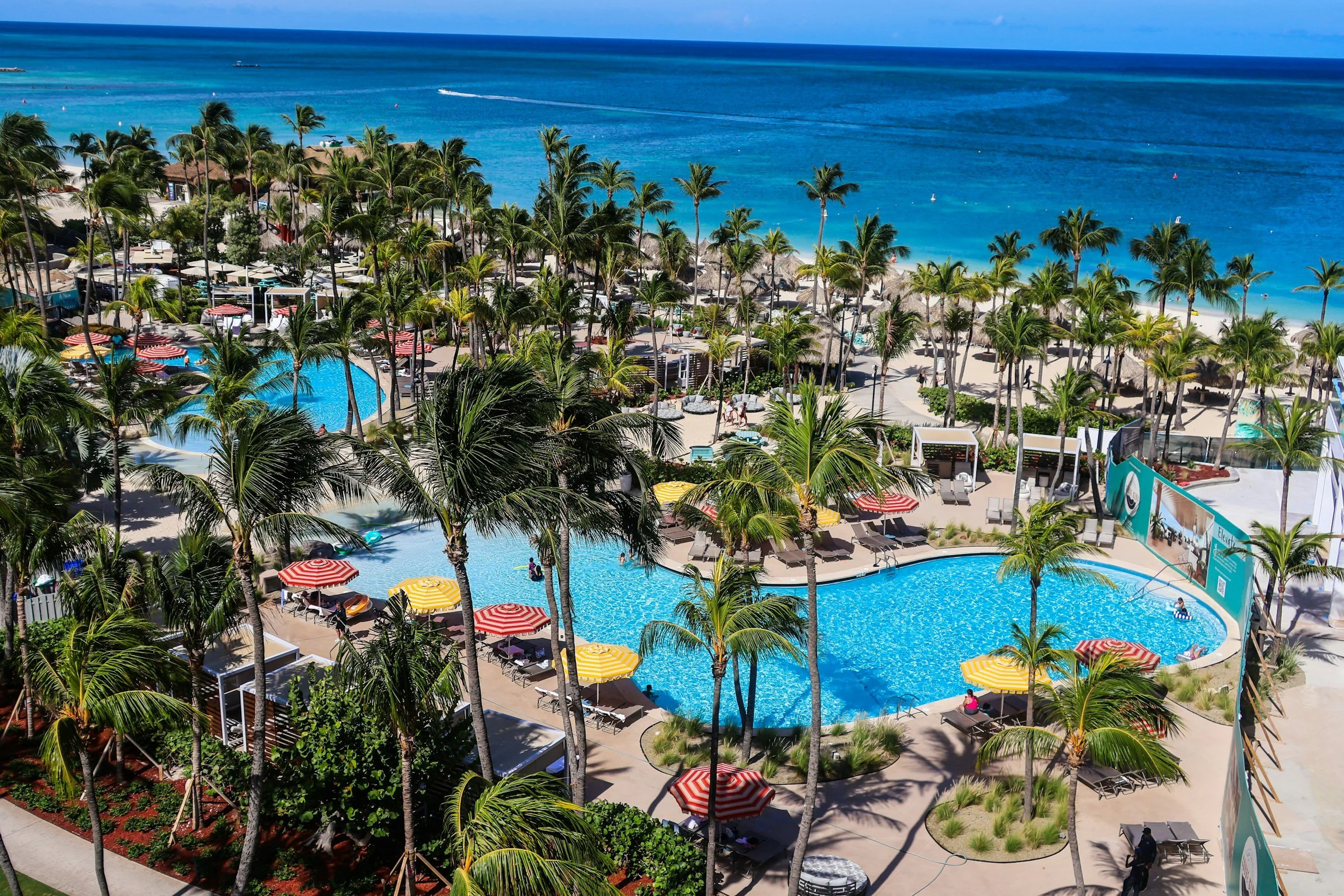 Aerial view of a tropical beach resort with palm trees, a large pool, colorful umbrellas, and the ocean in the background under a blue sky.