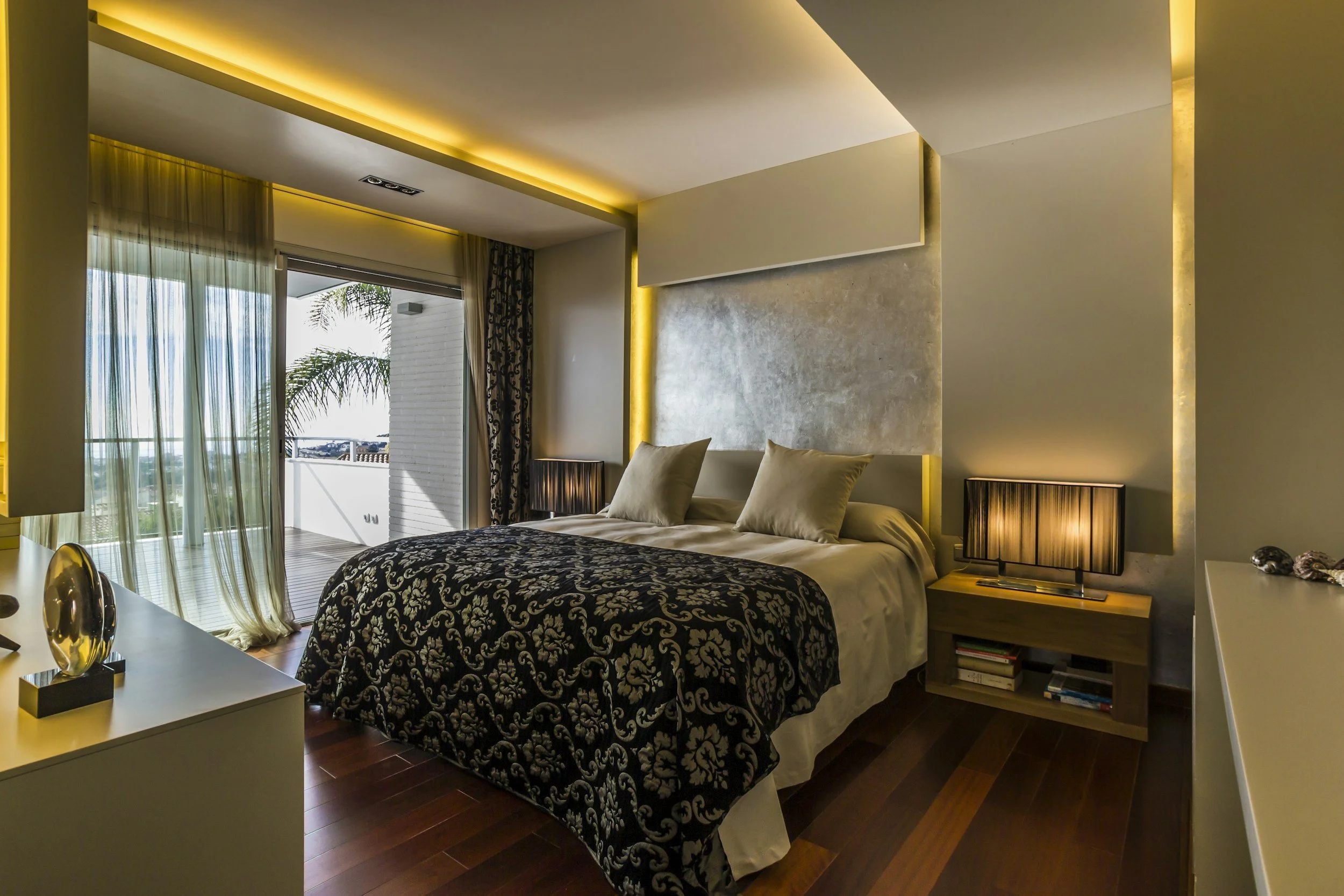 A modern bedroom with a bed, decorative pillows, nightstands with lamps, and large sliding glass doors leading to a balcony with a scenic view, illuminated by warm ambient lighting.