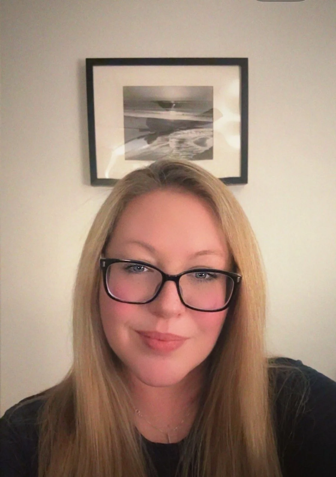 A woman with long blonde hair and glasses taking a selfie indoors, with a framed beach scene photograph on the wall behind her.