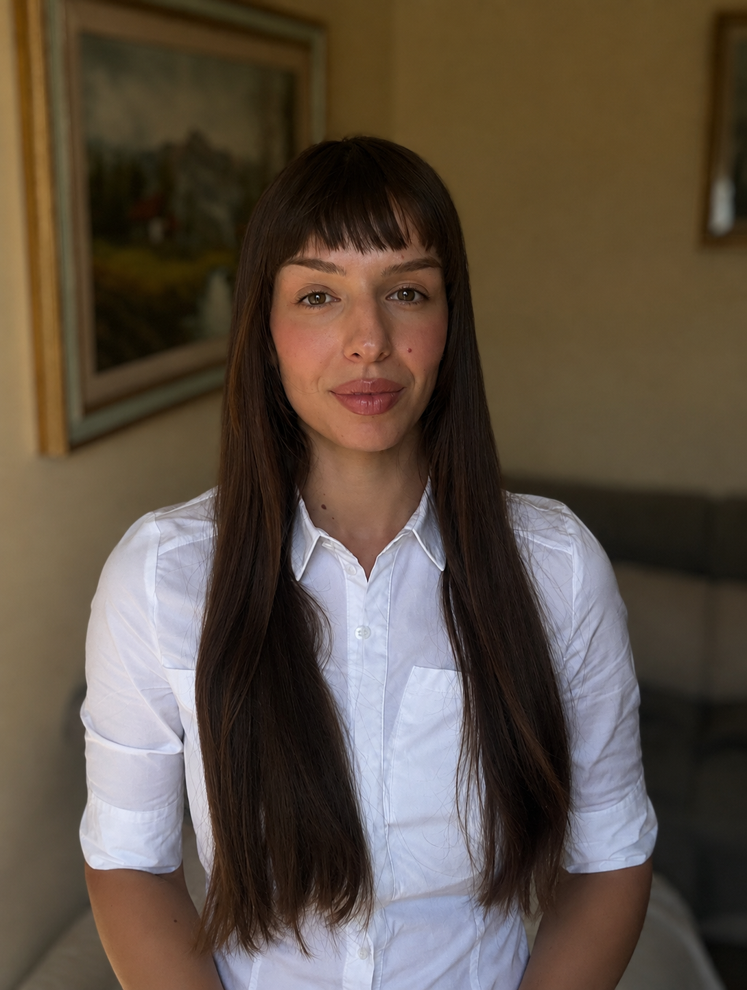 Psicologa con lunghi capelli castani e camicia bianca, posa in una stanza con dipinti sul muro.