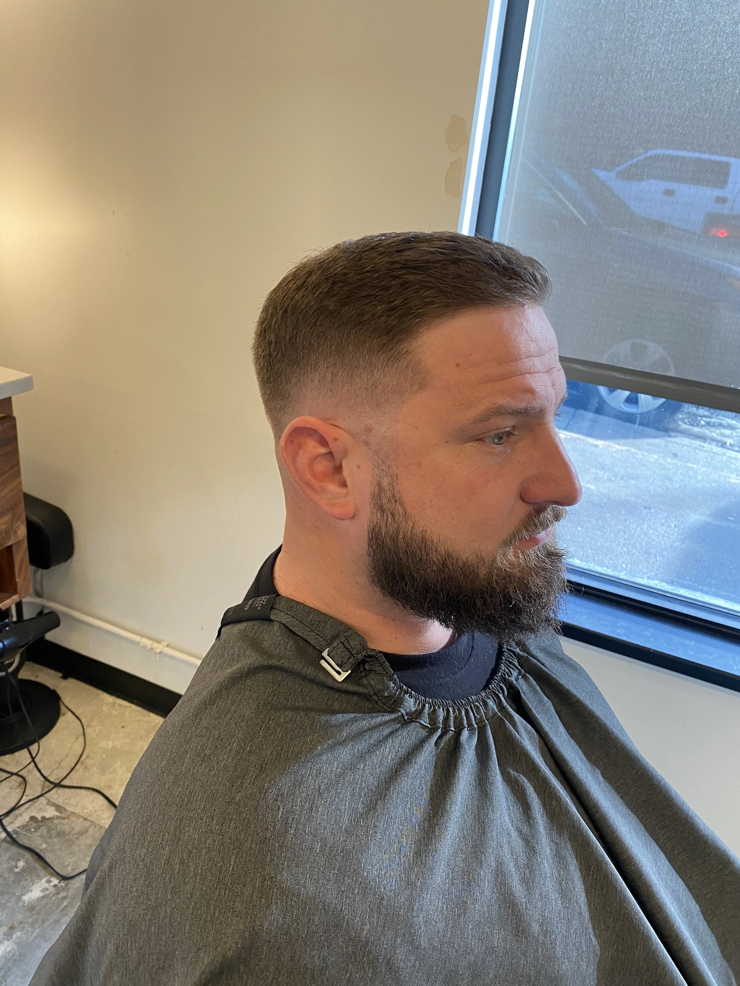 A man with a trimmed beard and brown hair sitting in a salon chair, covered with a black cape, looking out a window.