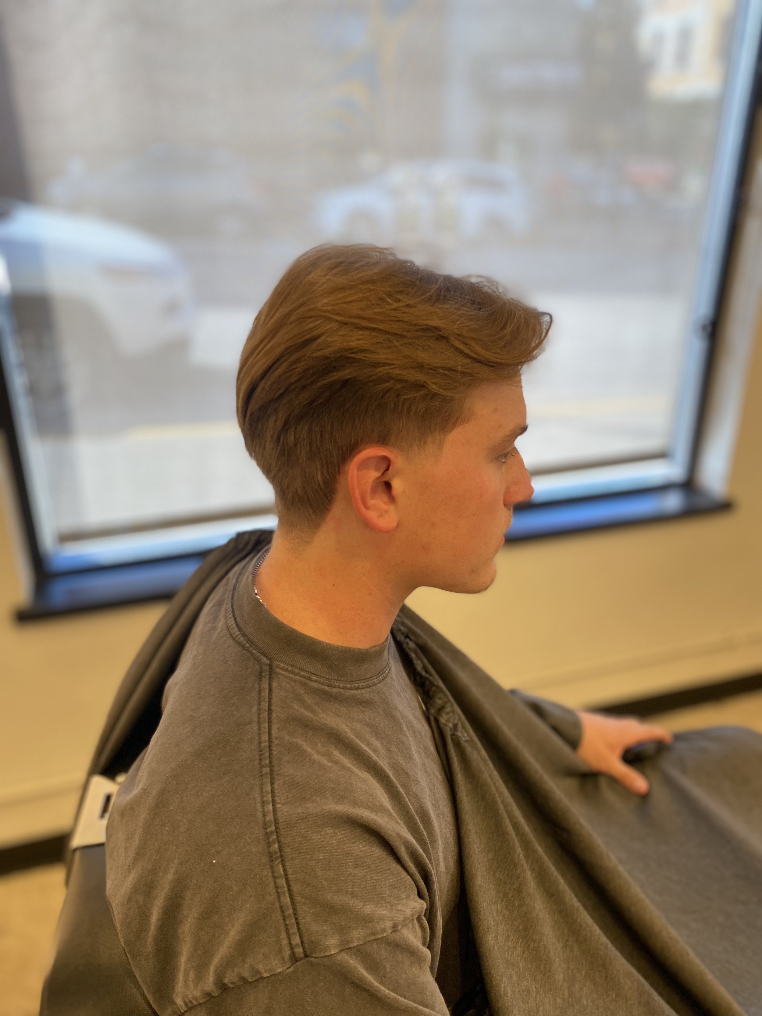 A young man with short, styled hair, sitting indoors near a window, wearing a gray sweatshirt, with a blurred view outside and a beige wall behind him.