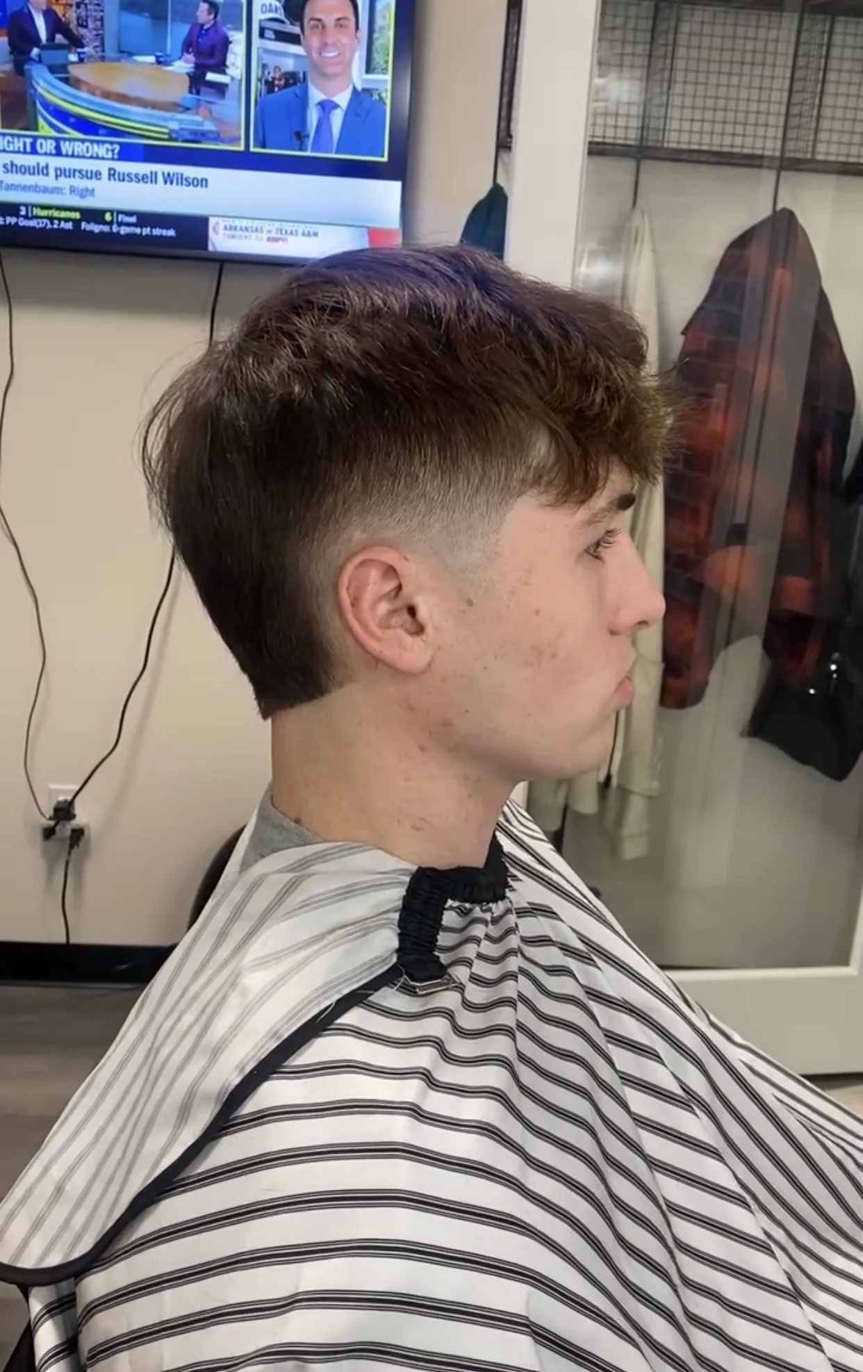 A young man with a fresh haircut, sitting in a barber's chair with a striped barber cape, in a barbershop, looking to the right. In the background, a television screen displays news anchors and a weather forecast.