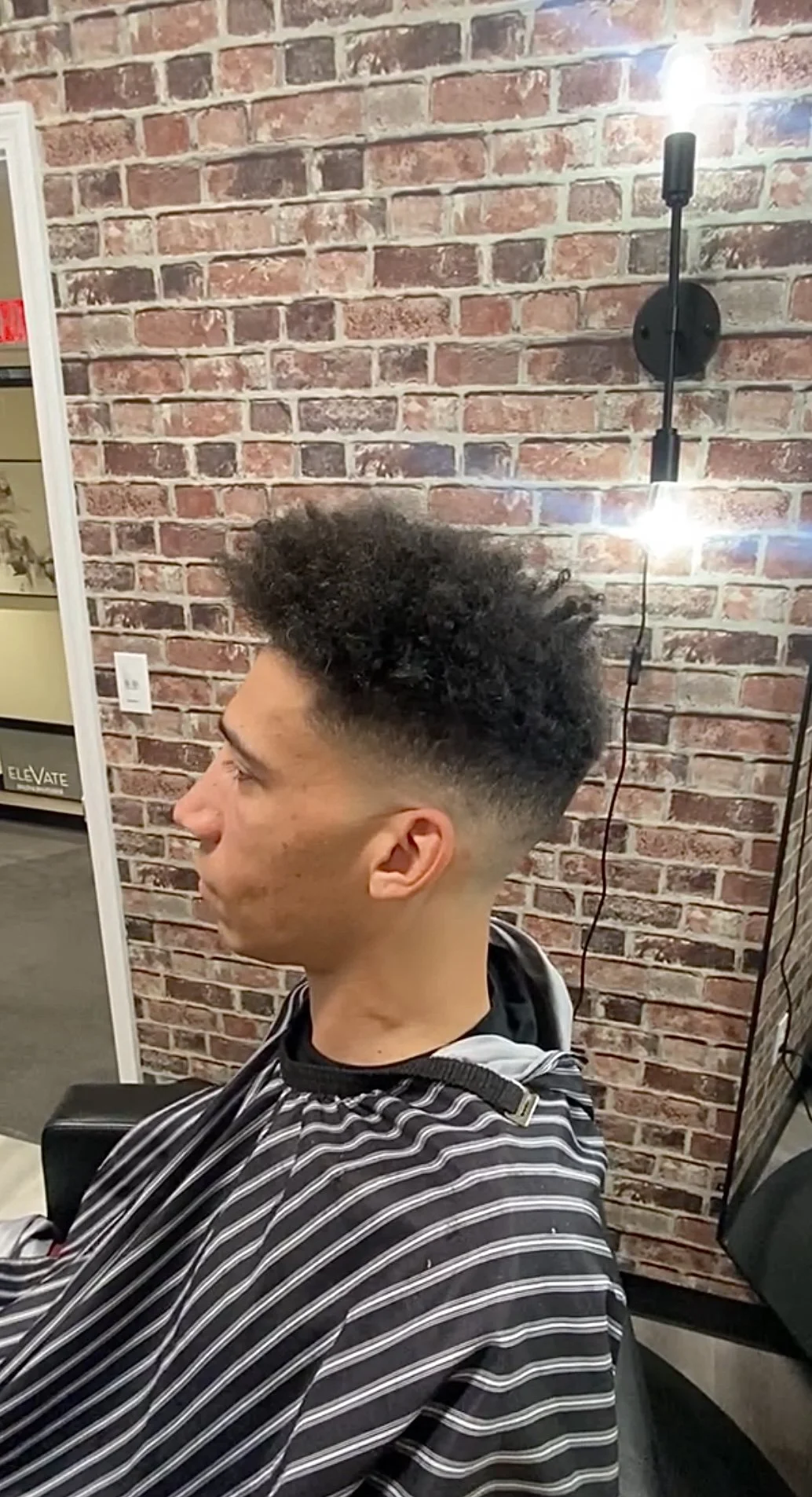 Profile of a young man with a fade hairstyle, sitting in a barber's chair, wearing a striped cape, against a red brick wall background.