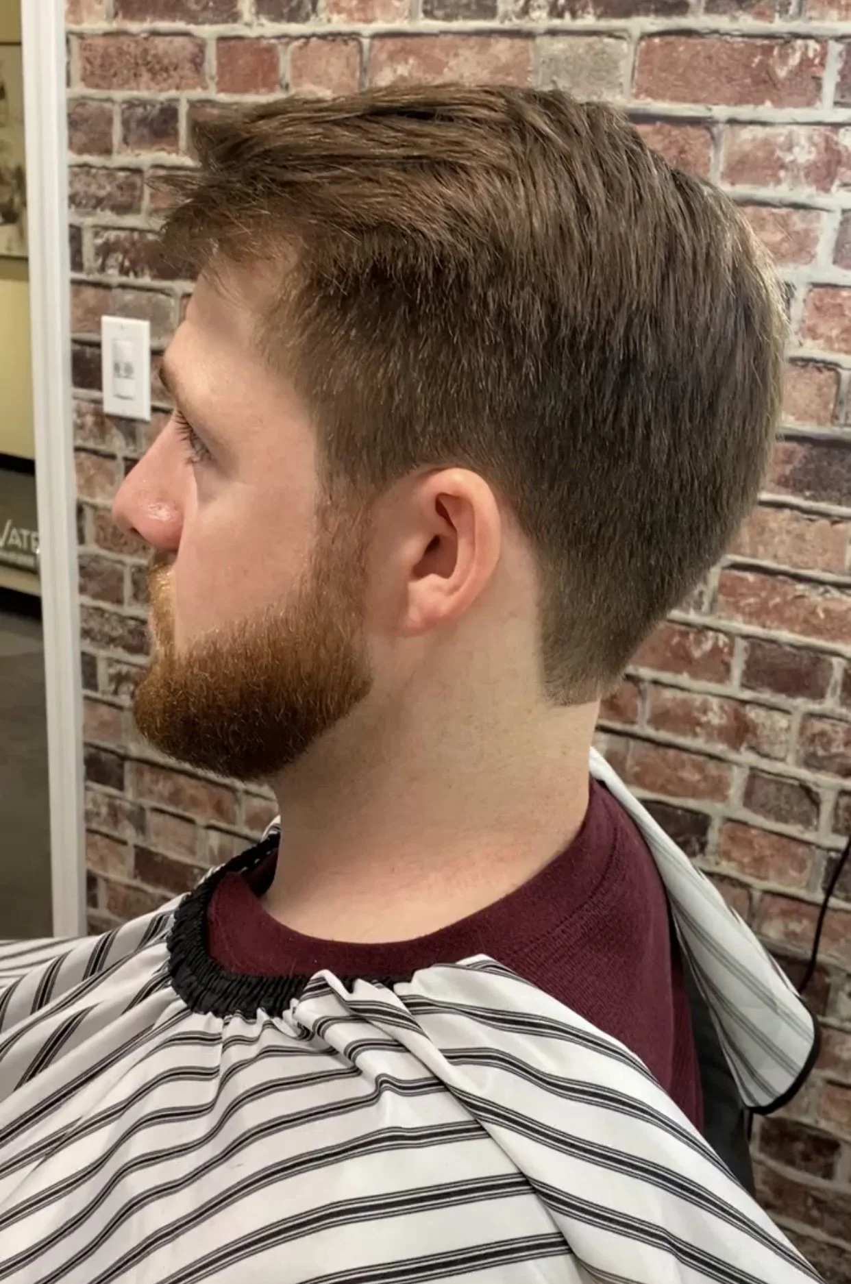 Side view of a young man with short, brown hair styled in a modern fade haircut, and a full reddish-brown beard, sitting in a barber's chair with a black and white striped cape around his shoulders, against a brick wall background.