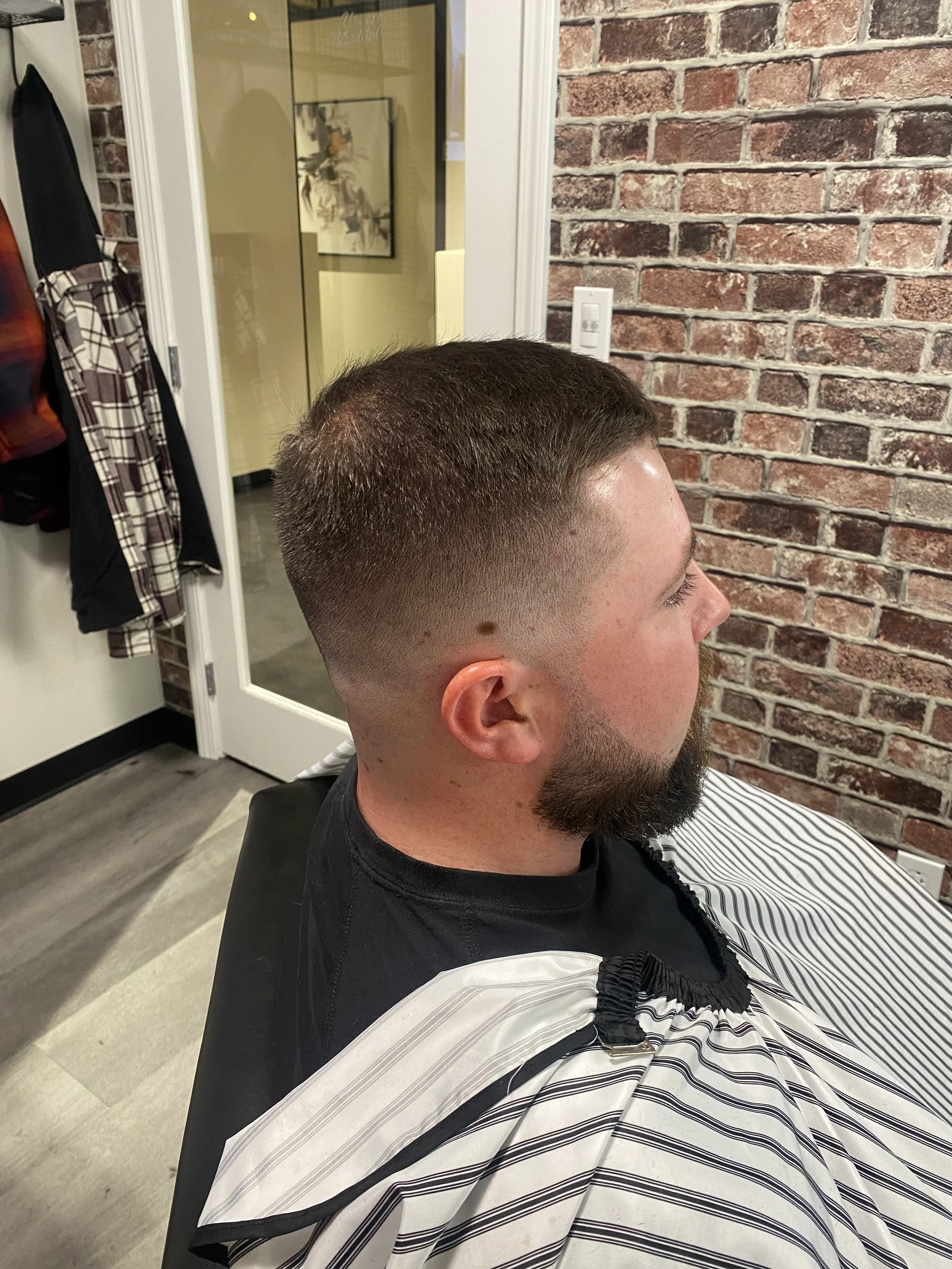 A man with dark hair and a beard sitting in a barber shop chair, wearing a barber cape with black and white stripes. The background features an exposed brick wall, a door with glass, and a mirror.