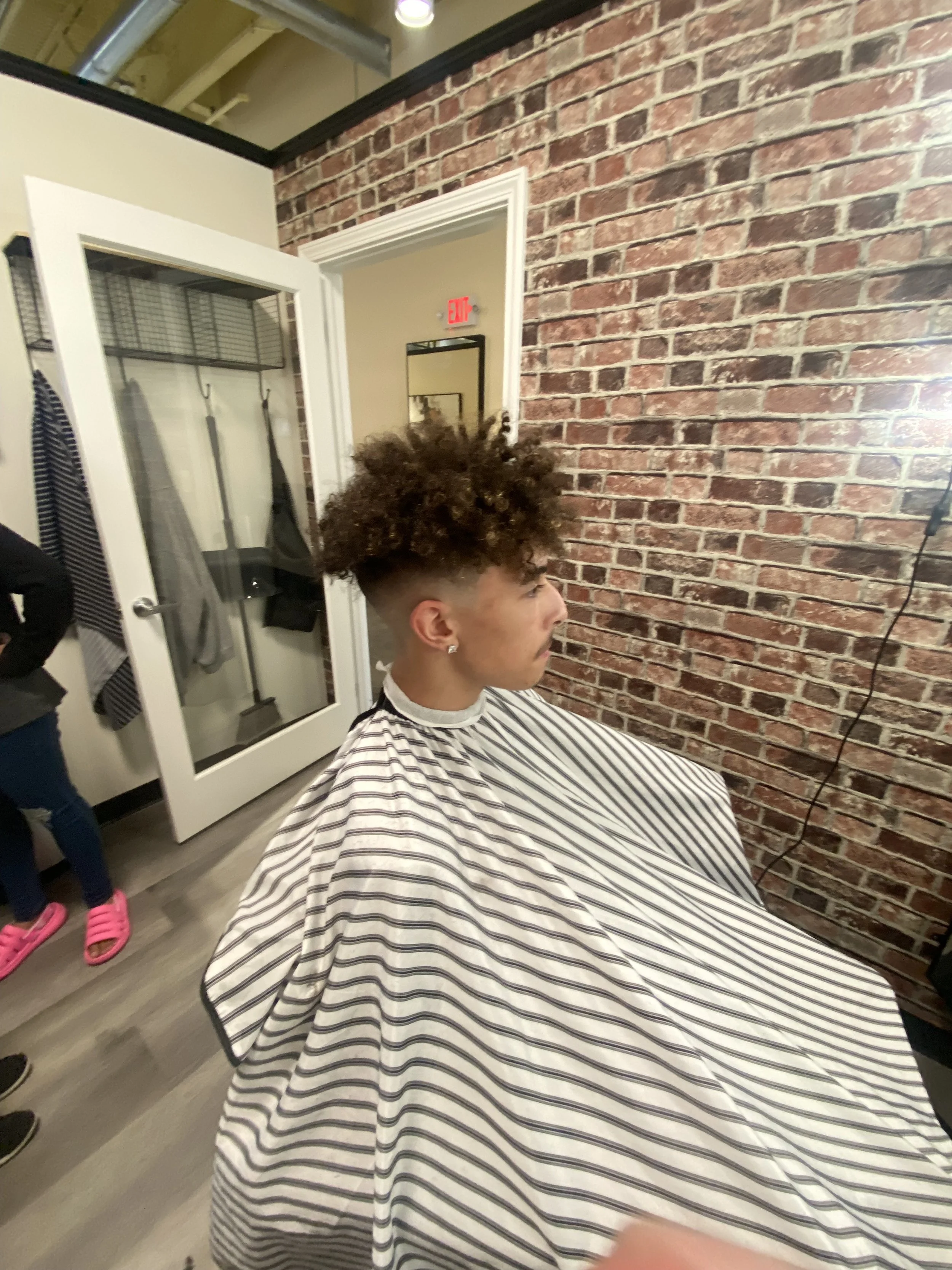 A young man with curly hair and earrings sitting in a barber's chair, wearing a striped barber's cape, inside a modern hair salon with an exposed brick wall.