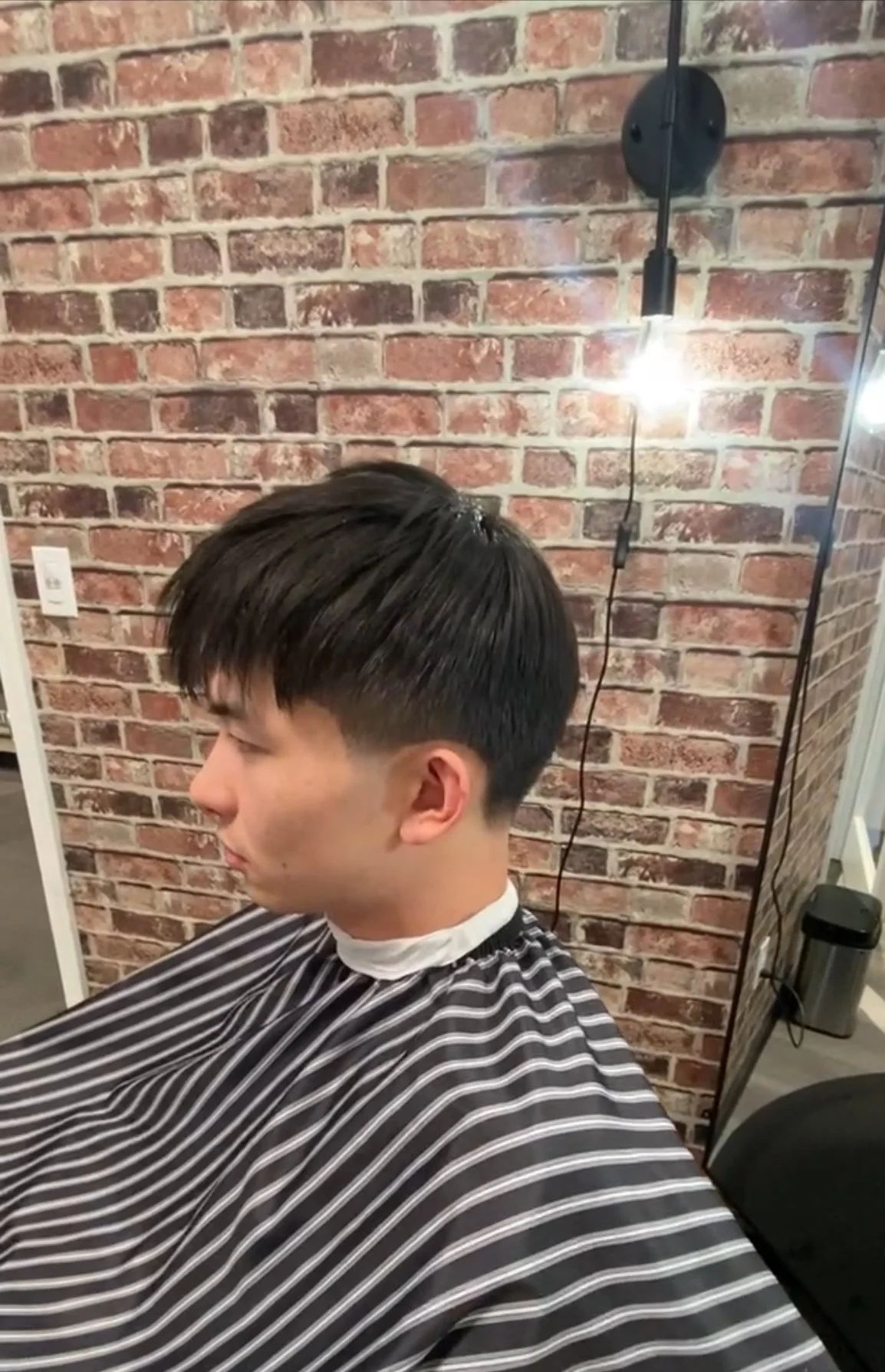 A young man getting a haircut in a salon with a brick wall backdrop. He is seated facing left, wearing a black and white striped barber cape, and has dark, freshly cut hair.