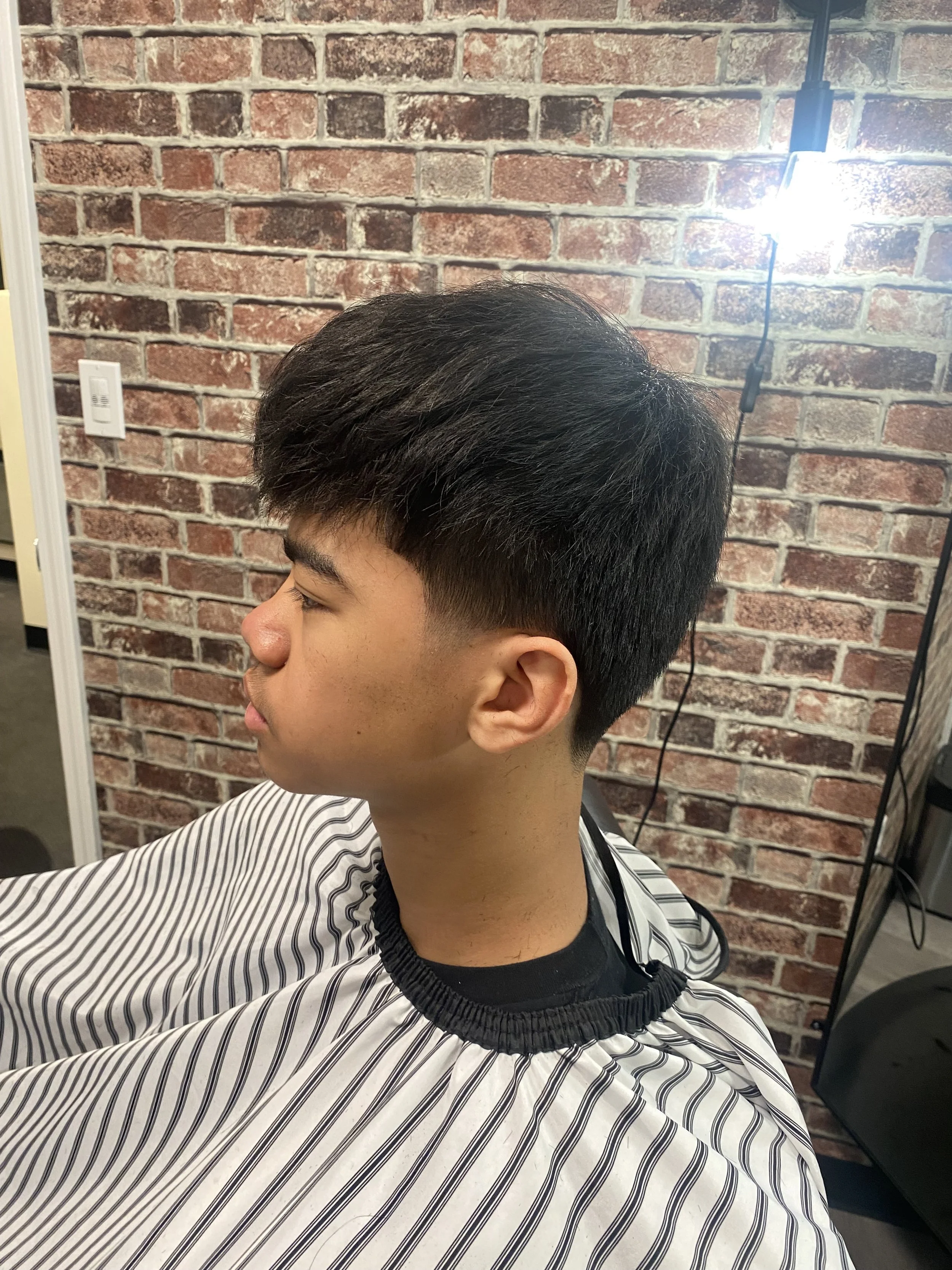 Young man with dark hair, wearing a striped barber cape, sitting in a barbershop chair, with a brick wall background and a bright salon light above.