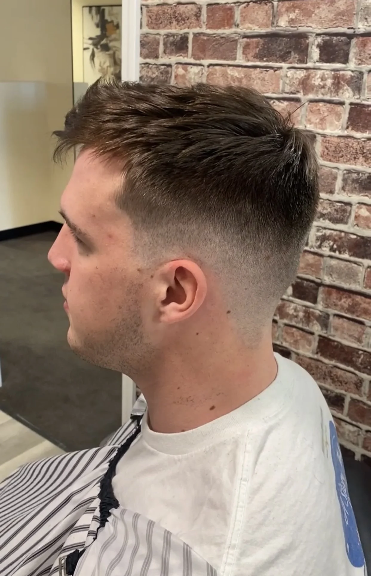 Young man with a modern undercut hairstyle sitting in a barber shop, facing left, against a brick wall background.