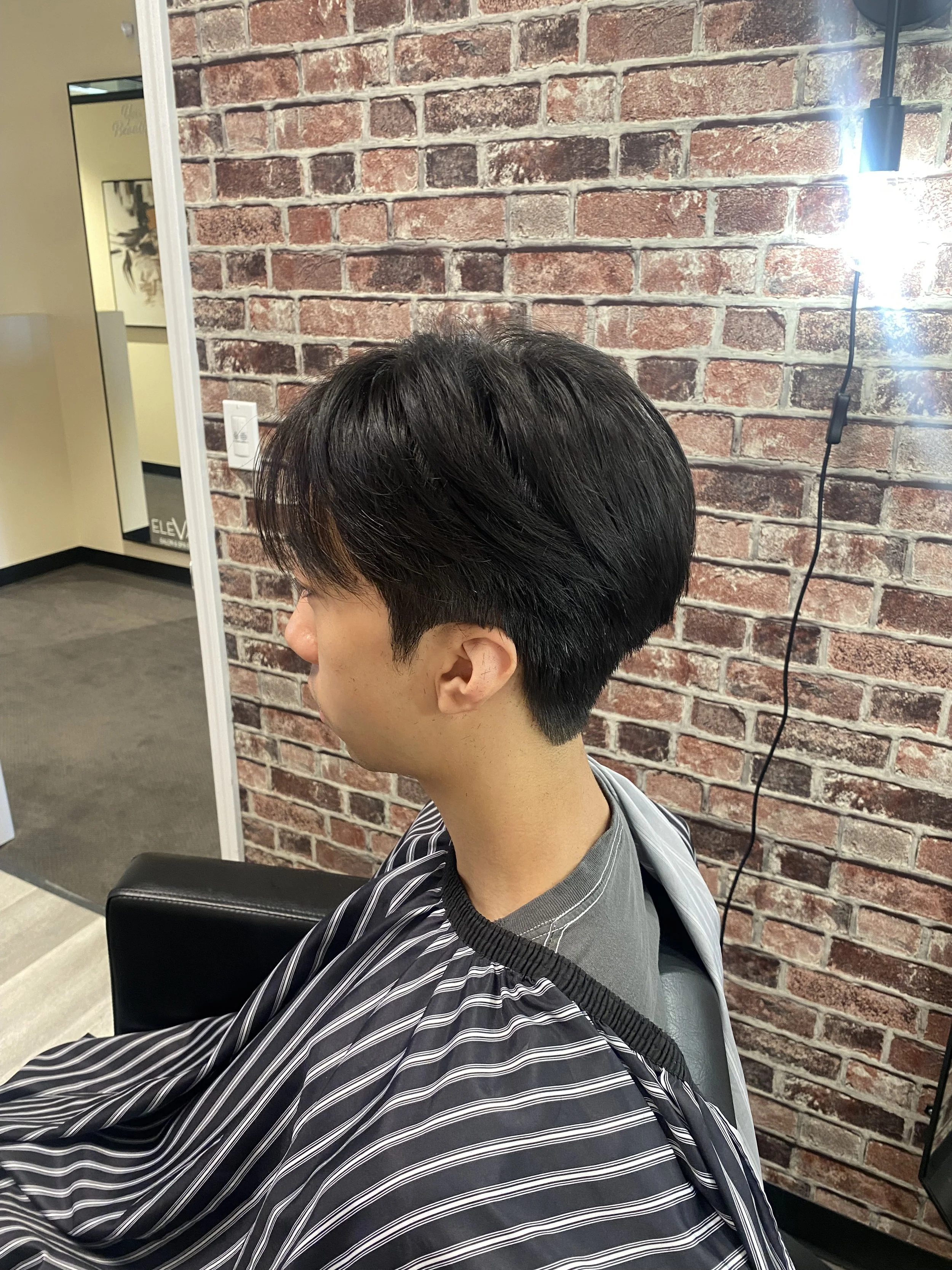 A person with short dark hair sitting in a salon chair wearing a black and white striped cape. A brick wall and a mirror are visible in the background.