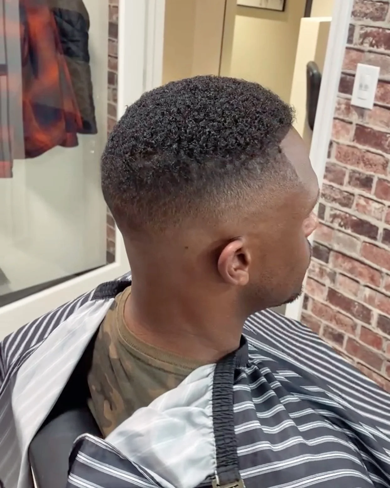 A man with a freshly cut hairstyle sitting in a barber's chair, with a fade haircut and textured top, in a barbershop with brick wall decor.