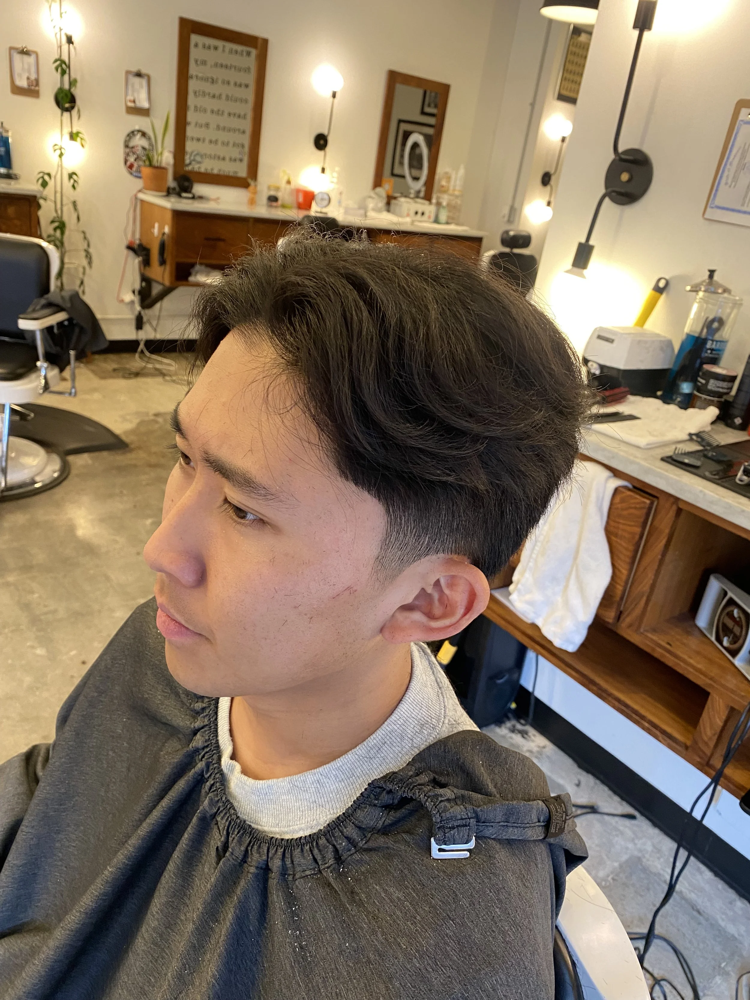 Young man with dark hair, sitting in a salon chair, getting a haircut, with mirrors and salon equipment visible in the background.