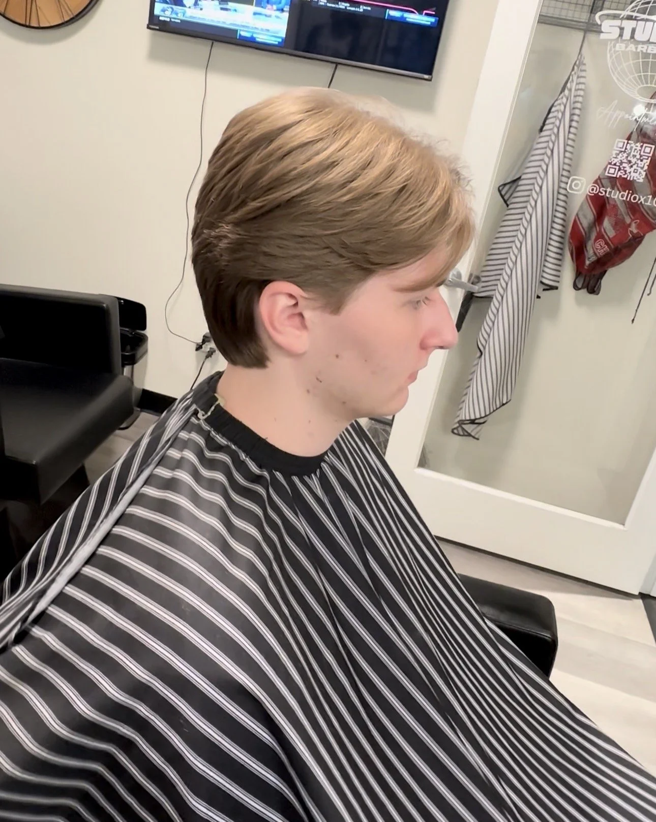 A young man with light brown hair sitting in a barber's chair in a hair salon, wearing a black and white striped cape. Behind him, a glass door with a towel and a red bag hanging on it, and a wall-mounted TV screen showing a computer desktop.