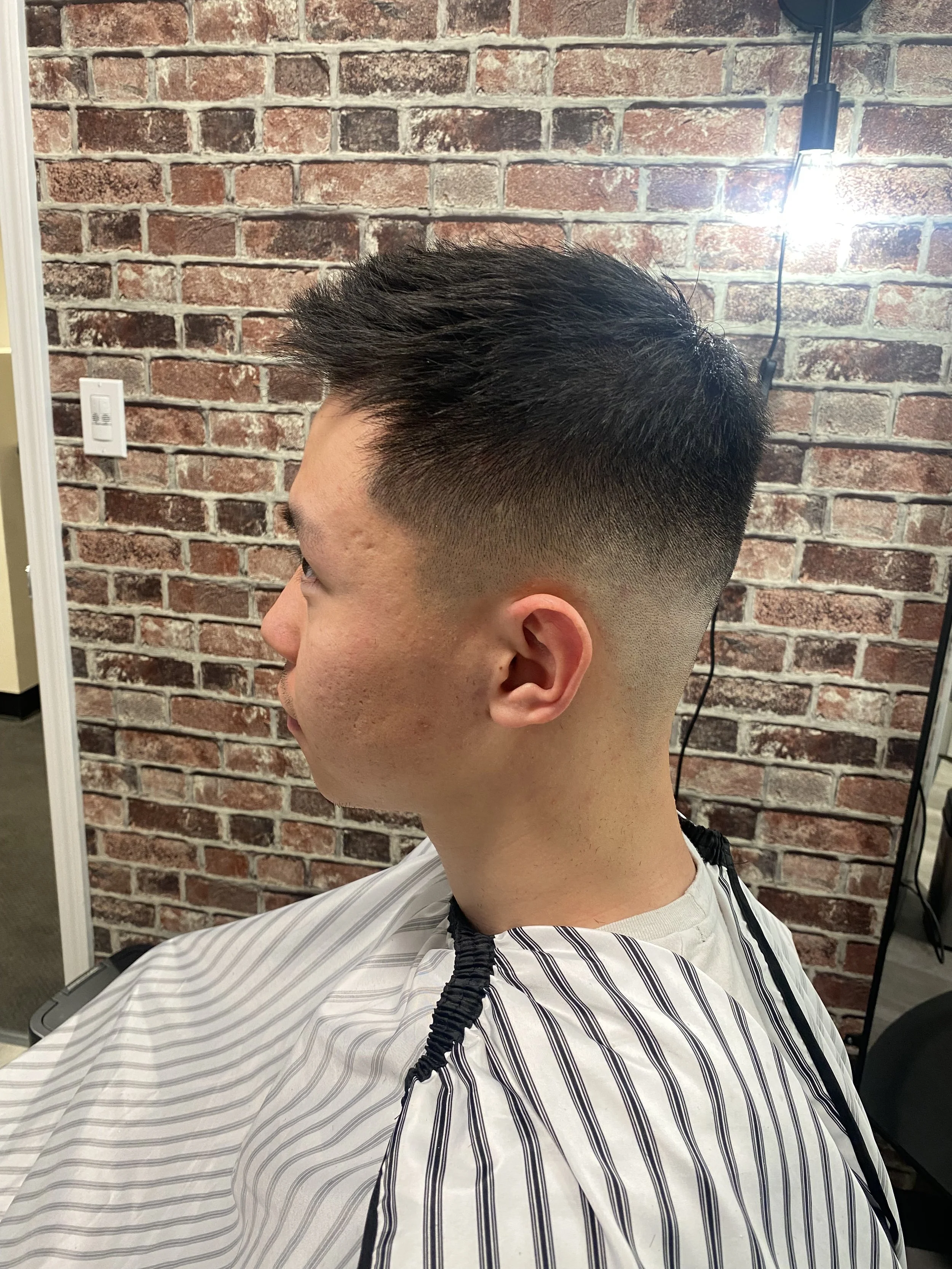 A man with a short, styled haircut sitting in a barber shop chair wearing a barber cape with black and white stripes, against a brick wall background.
